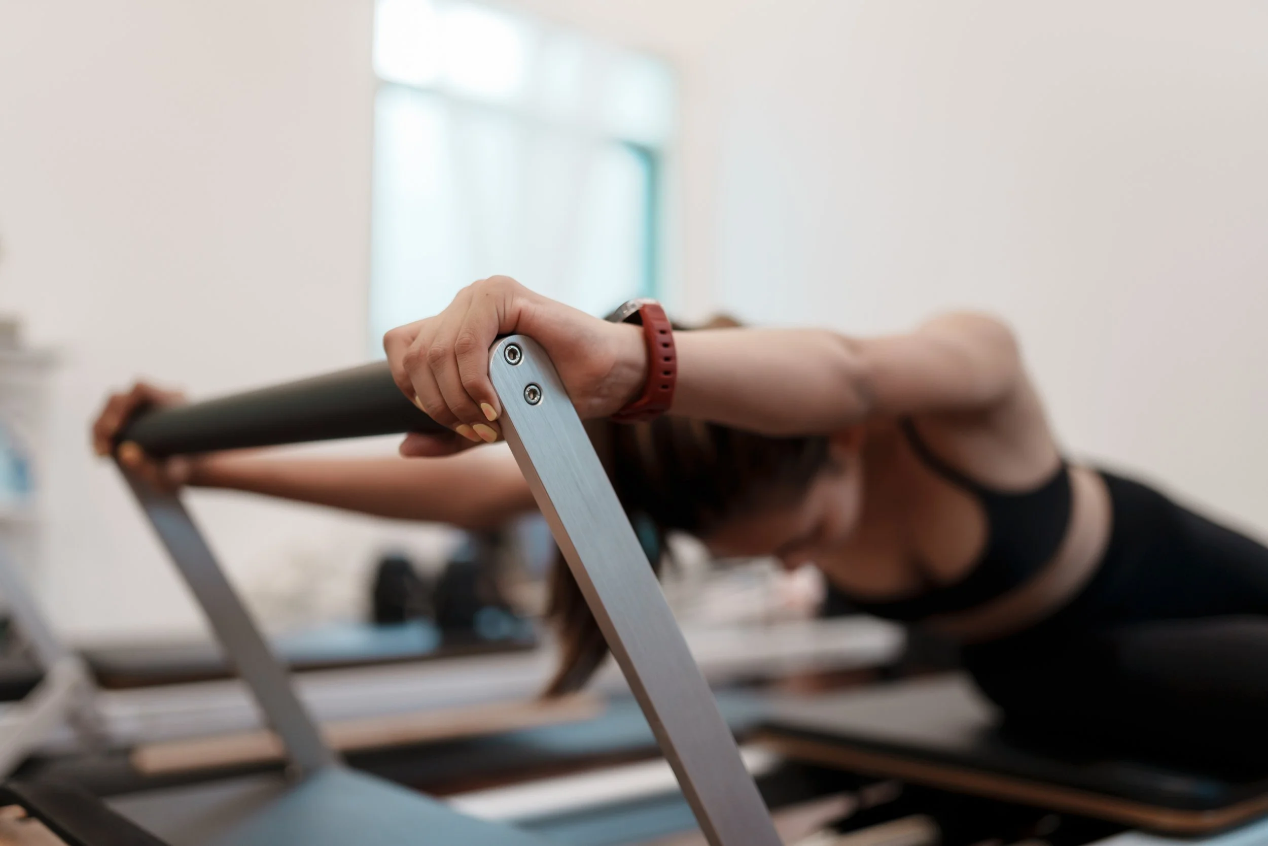Person lying face down on a reformer with arms extended forward, holding a barbell during a workout in a gym.
