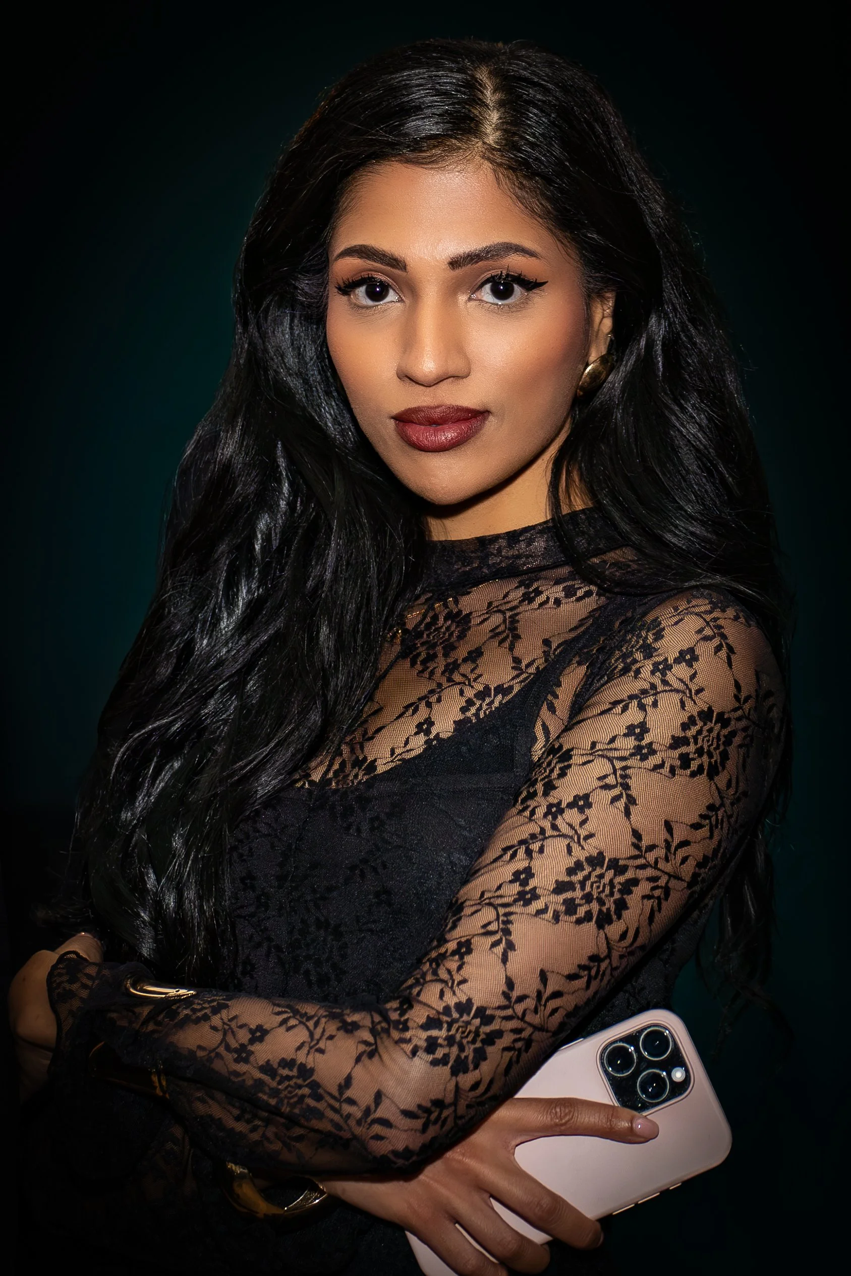 A woman with long black hair holding an iPhone, wearing a black lace top with floral patterns on the sleeves, against a dark background.