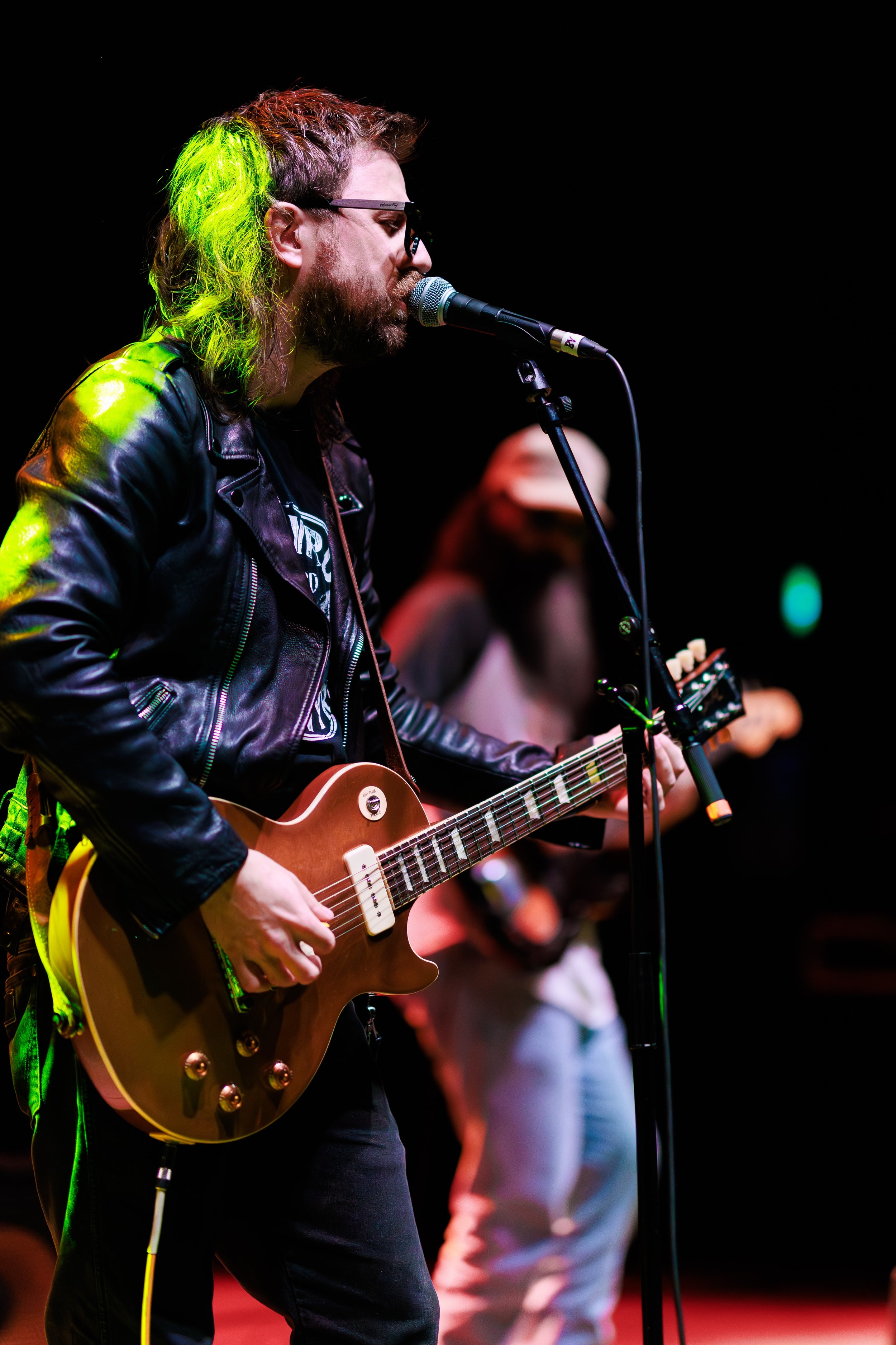 A musician with long hair, glasses, and a beard, wearing a black leather jacket, is playing an acoustic guitar and singing into a microphone on stage with colorful lighting. In the background, another person can be seen holding a percussion instrumen