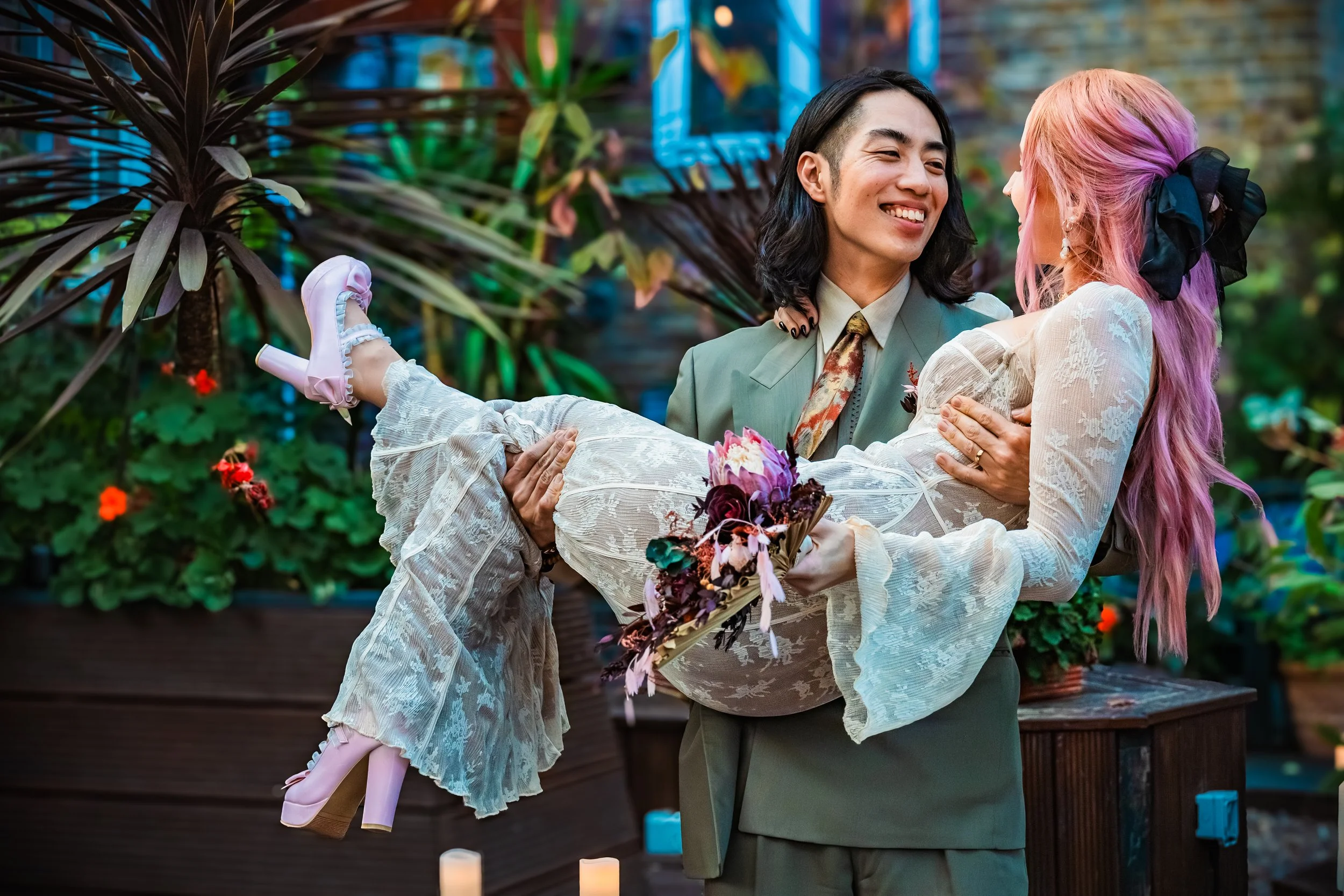 A man with long black hair in a suit is smiling while holding a woman with pink hair dressed in white lace and pink heels, who is holding a bouquet of flowers, in a lush, colorful garden with plants and candles.