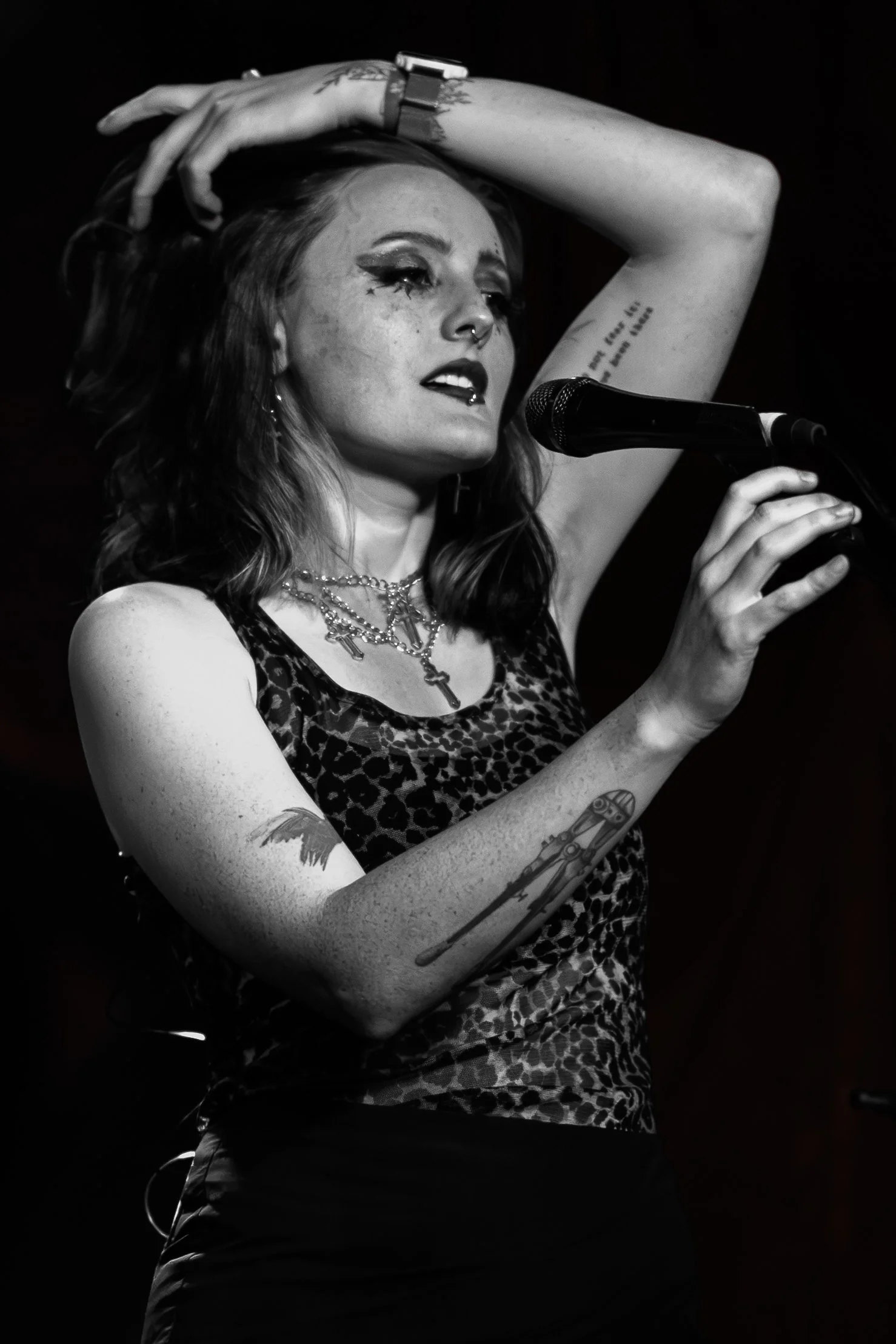 A woman with tattoos on her arms, wearing a leopard print top, and necklaces, standing in front of a microphone on stage, with her eyes closed and one hand raised above her head.