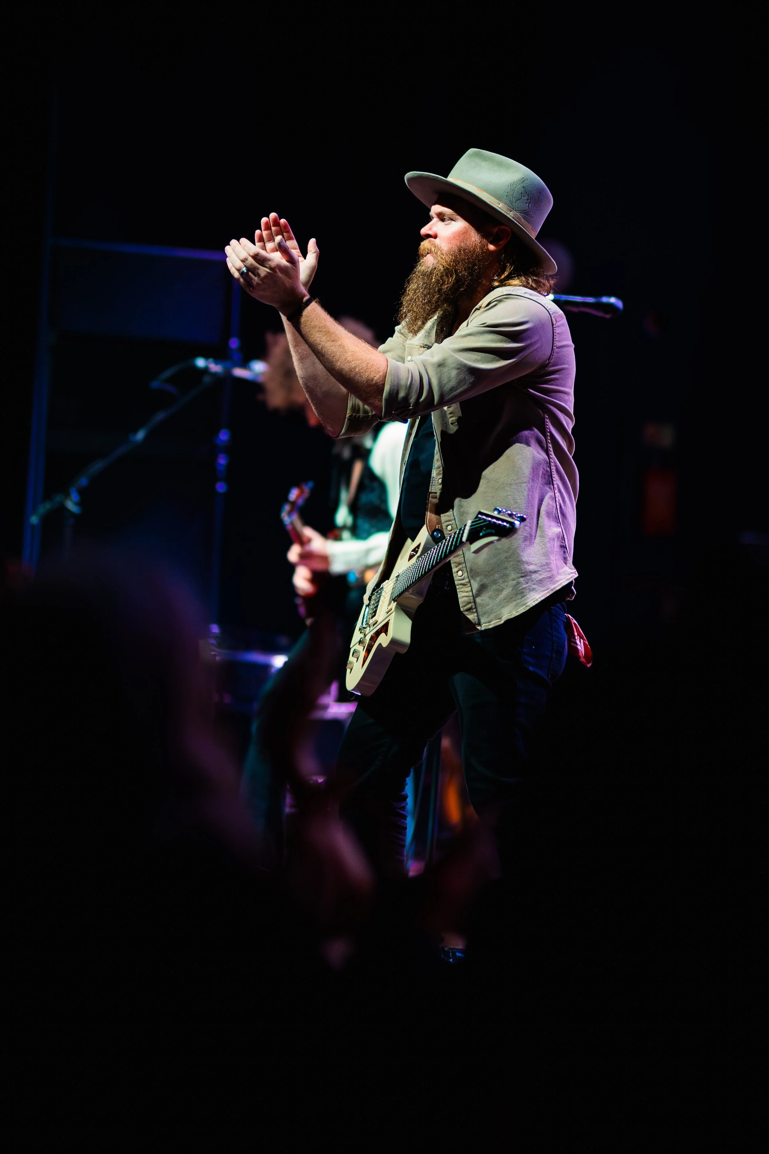 A musician with a beard and long hair wearing a wide-brimmed hat, beige jacket, and playing an electric guitar during a live performance on stage with dark background and concert lighting.