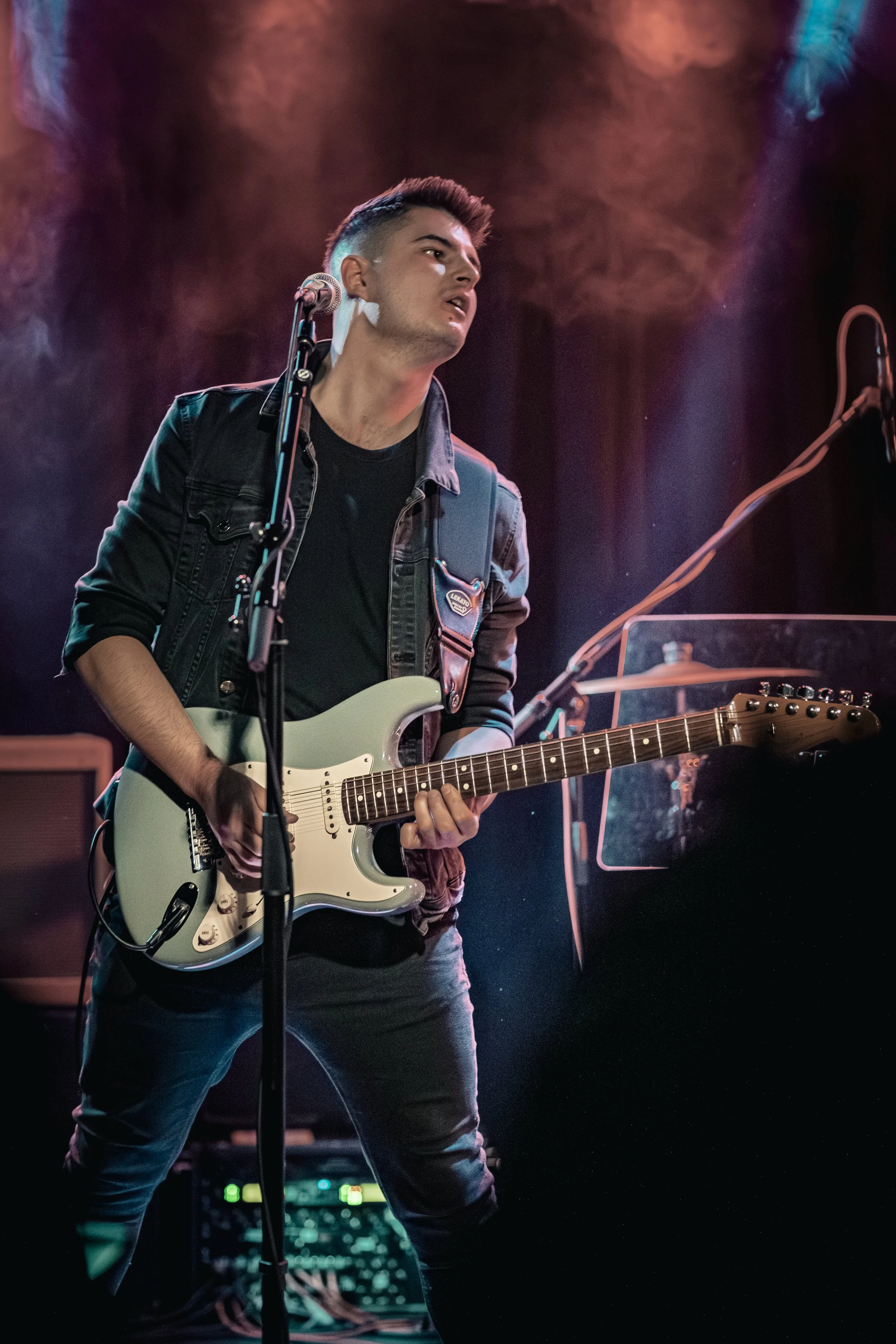 A young man performing with a white electric guitar on stage, wearing a black jacket and black T-shirt, with colorful stage lights and fog effects.