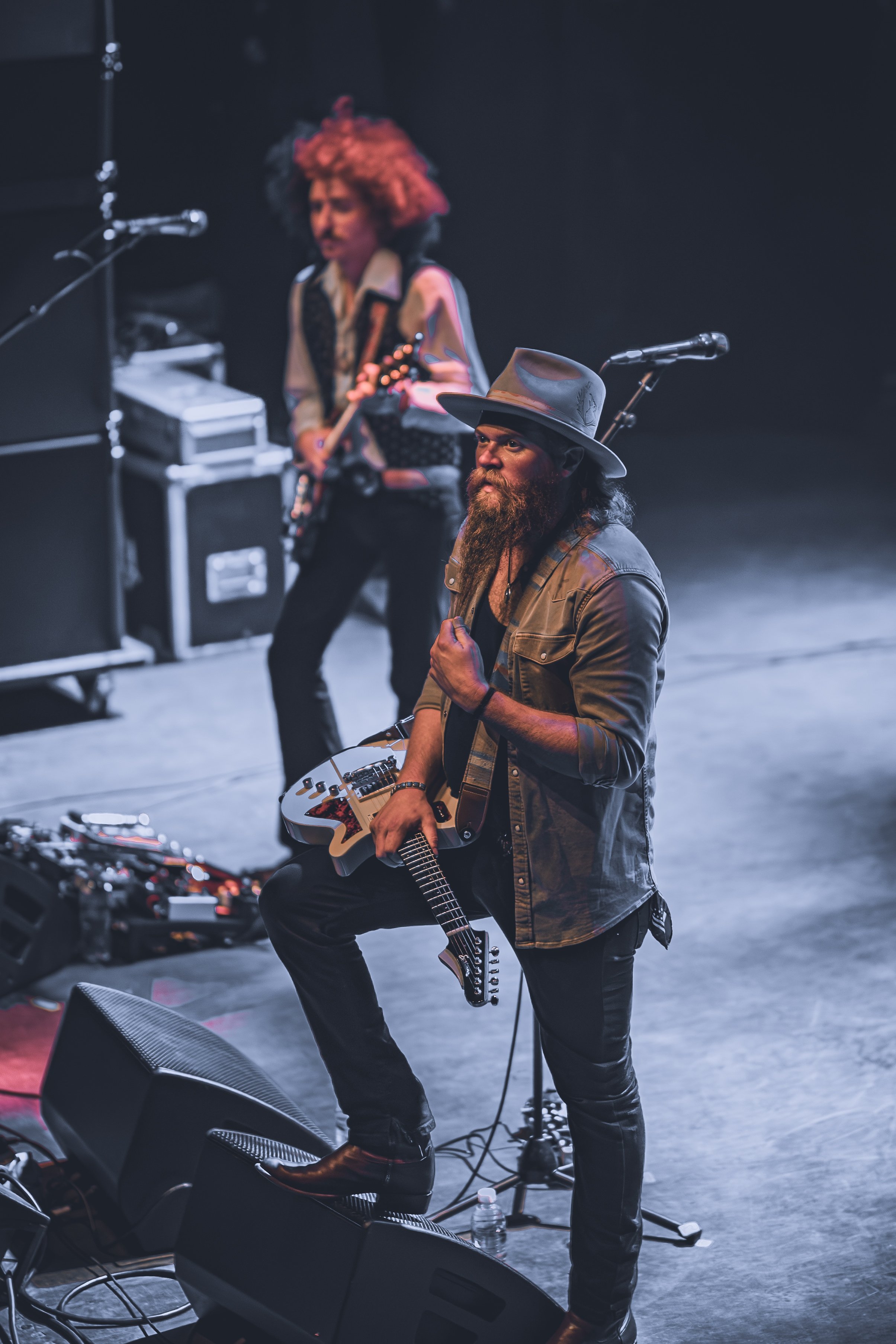 Two musicians performing on stage. The male musician in the foreground has a full beard, long hair, and is wearing a wide-brimmed hat, a denim jacket, and black pants. He is playing an electric guitar and kneeling on one knee. The female musician in 