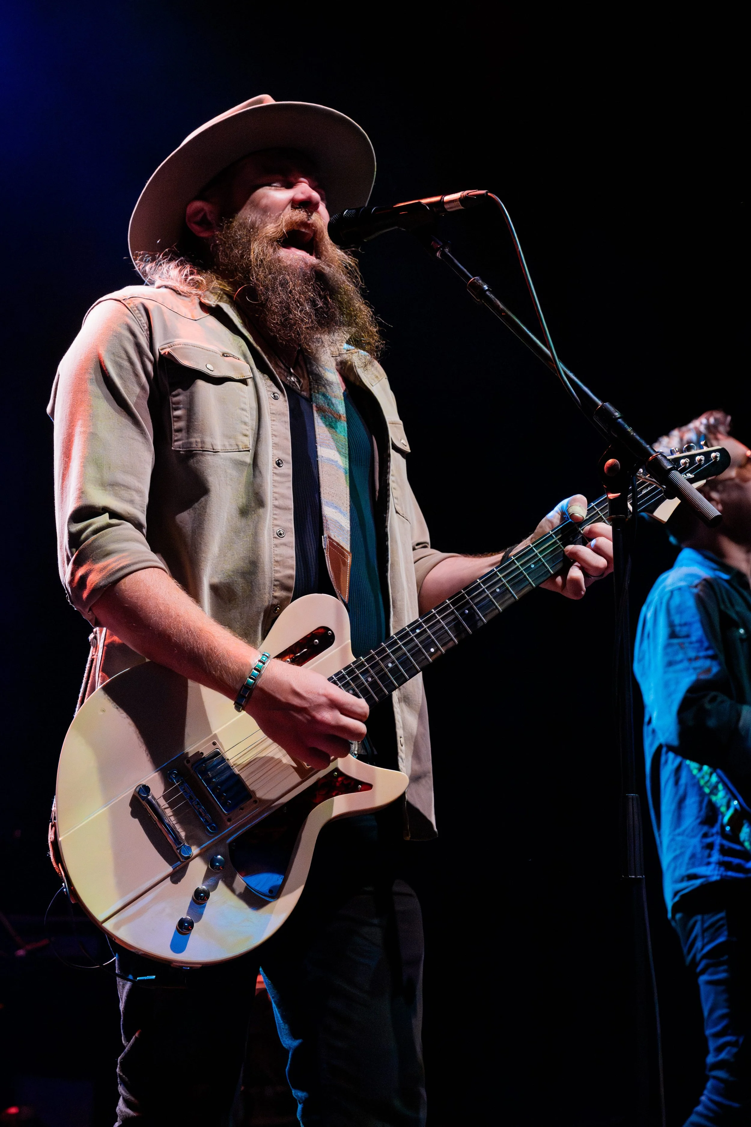 Man with beard wearing a cowboy hat singing into a microphone while playing an electric guitar on stage at a concert.