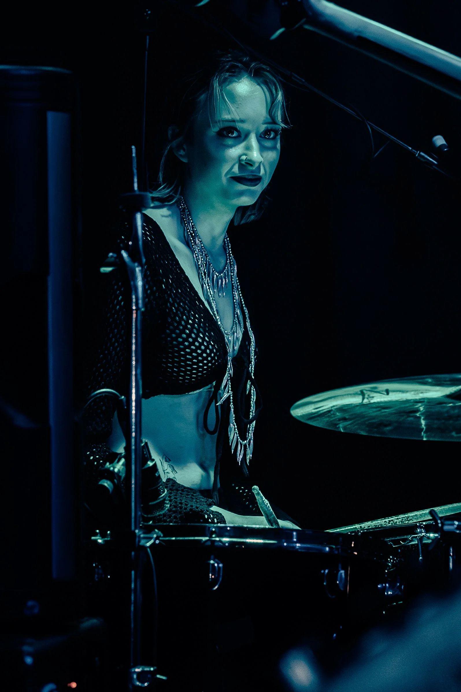A woman with short blonde hair and dark makeup, wearing a black fishnet top with jewelry, is playing drums in a dimly lit setting.