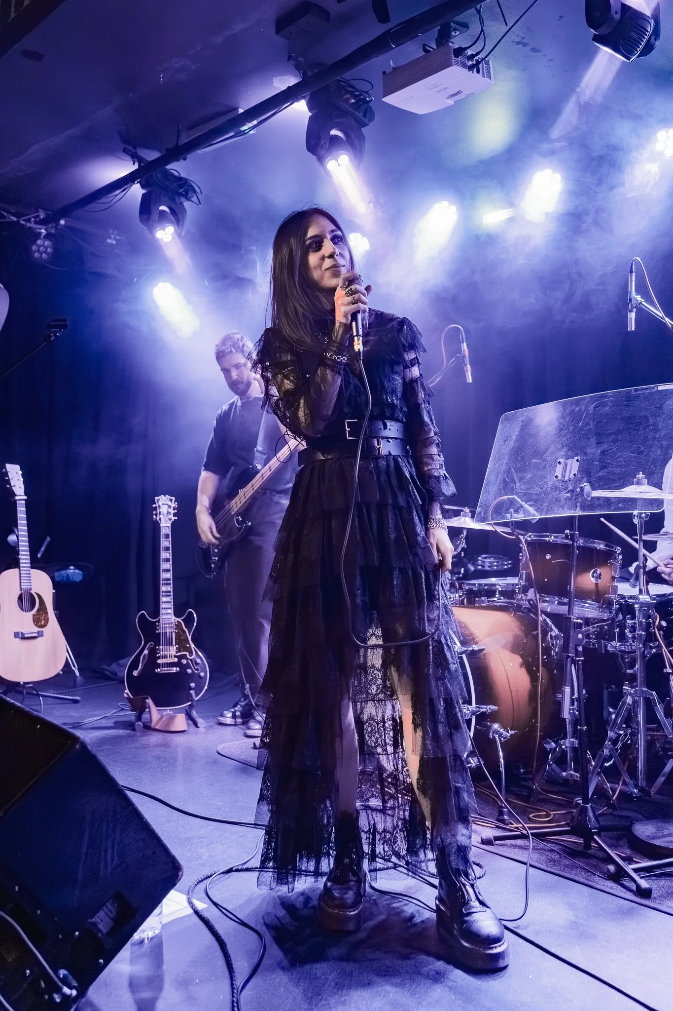 Female singer in a black, lace dress and platform boots performing on stage, with band members and musical instruments including guitars, drums, and microphones visible under stage lighting.