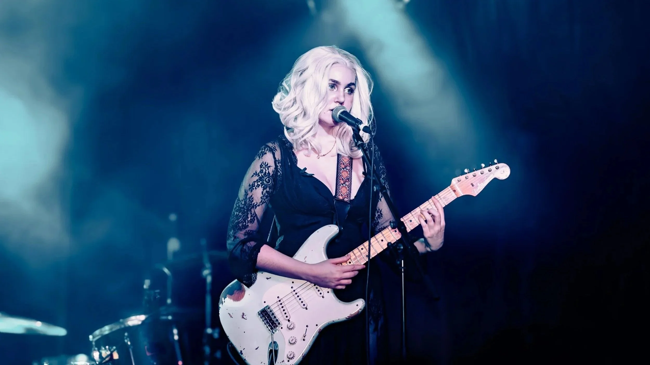 A female musician with blonde hair playing an electric guitar on stage.