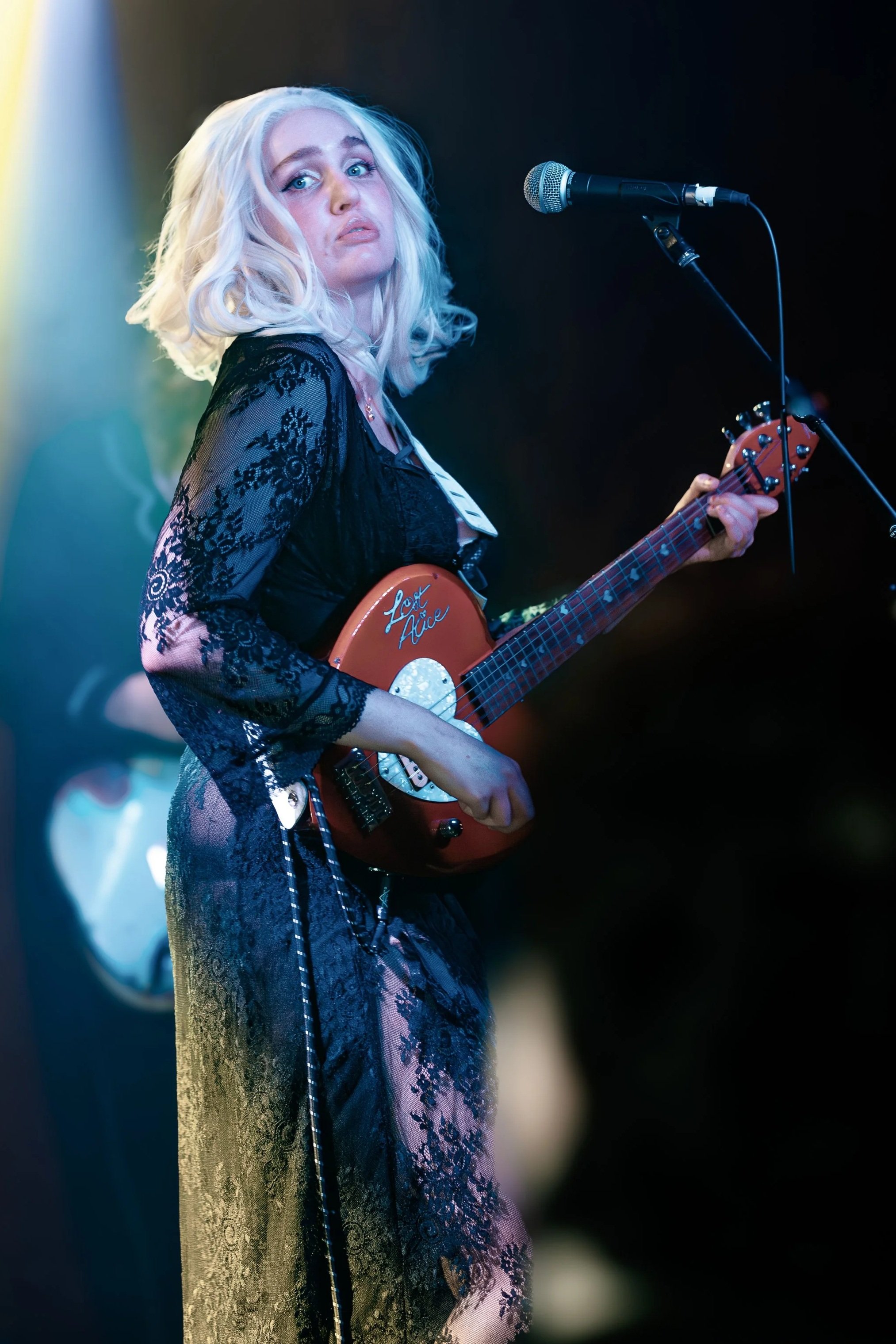 A woman with platinum blonde hair plays guitar on stage, illuminated by yellow and blue stage lights, wearing a black lace dress.