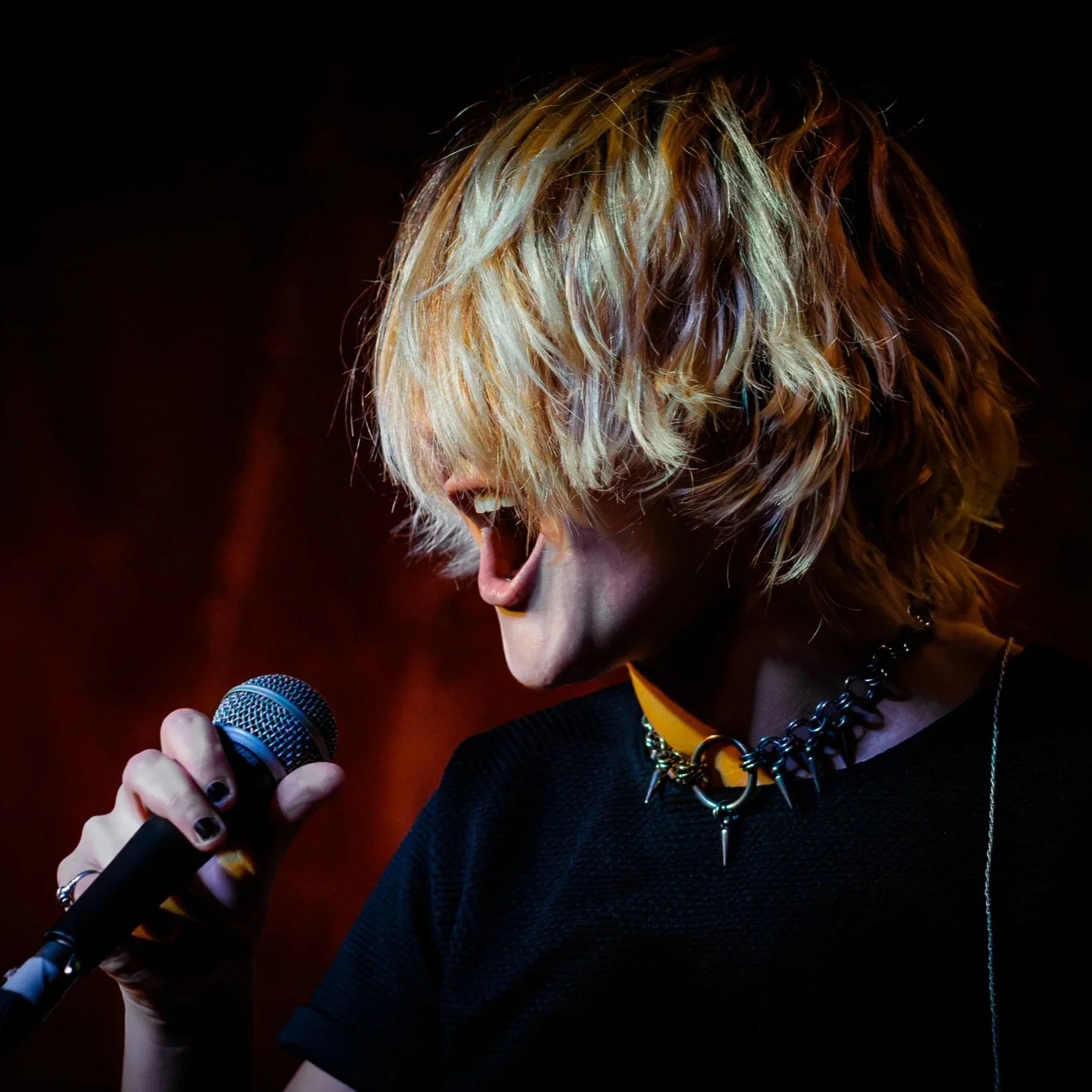 A person with blonde, tousled hair singing into a microphone on stage, wearing a black shirt and spiked choker necklace.