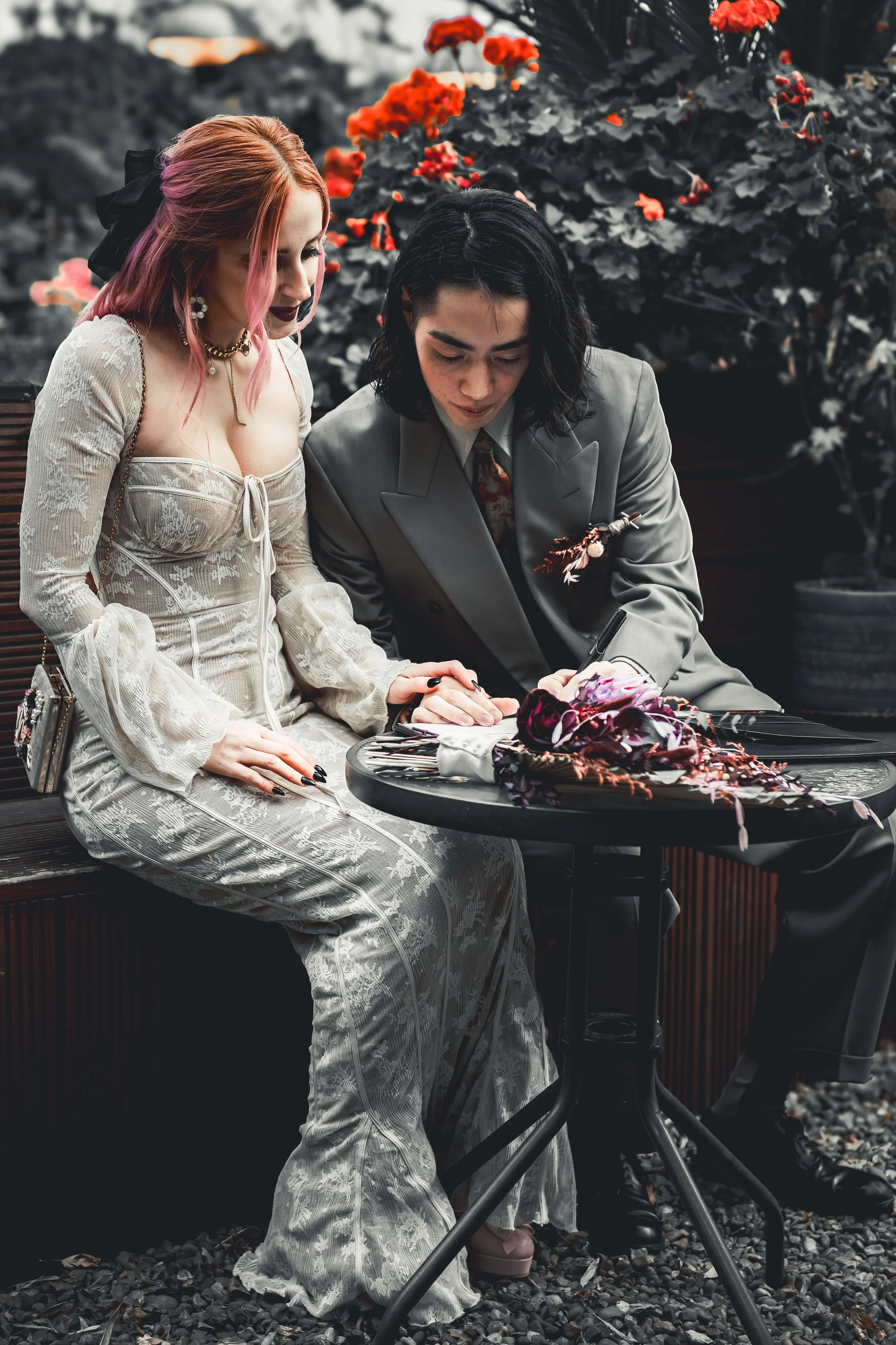 A woman with pink hair wearing a vintage lace dress sitting next to a man with black hair in a suit, both looking at and signing a book or magazine on a small round table with flowers. They are outdoors among vibrant red and white flowers.