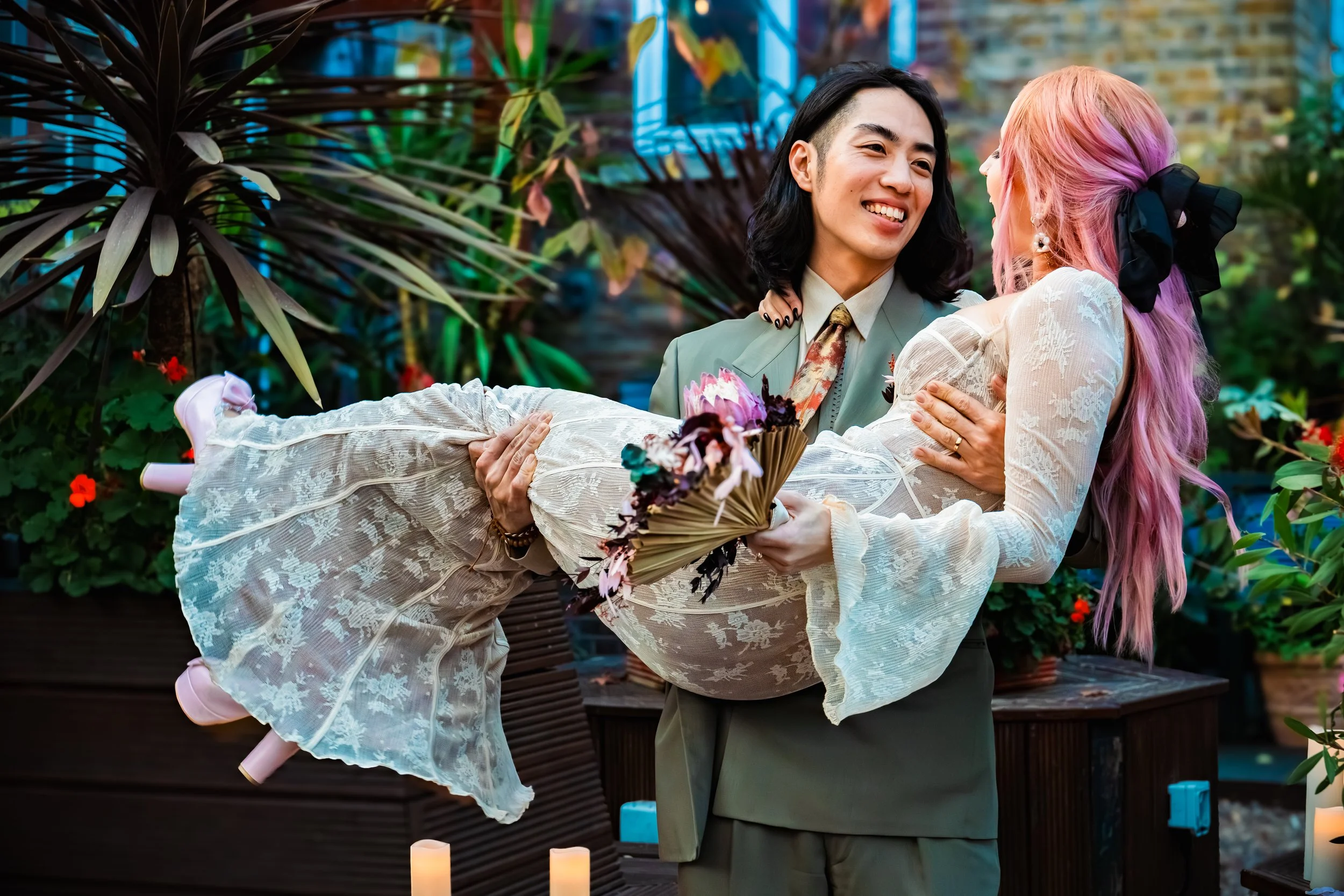 A man in a green suit smiling as he holds a woman with pink and purple hair in a cream-colored lace dress, who is holding a bouquet of flowers, in a lush indoor garden.