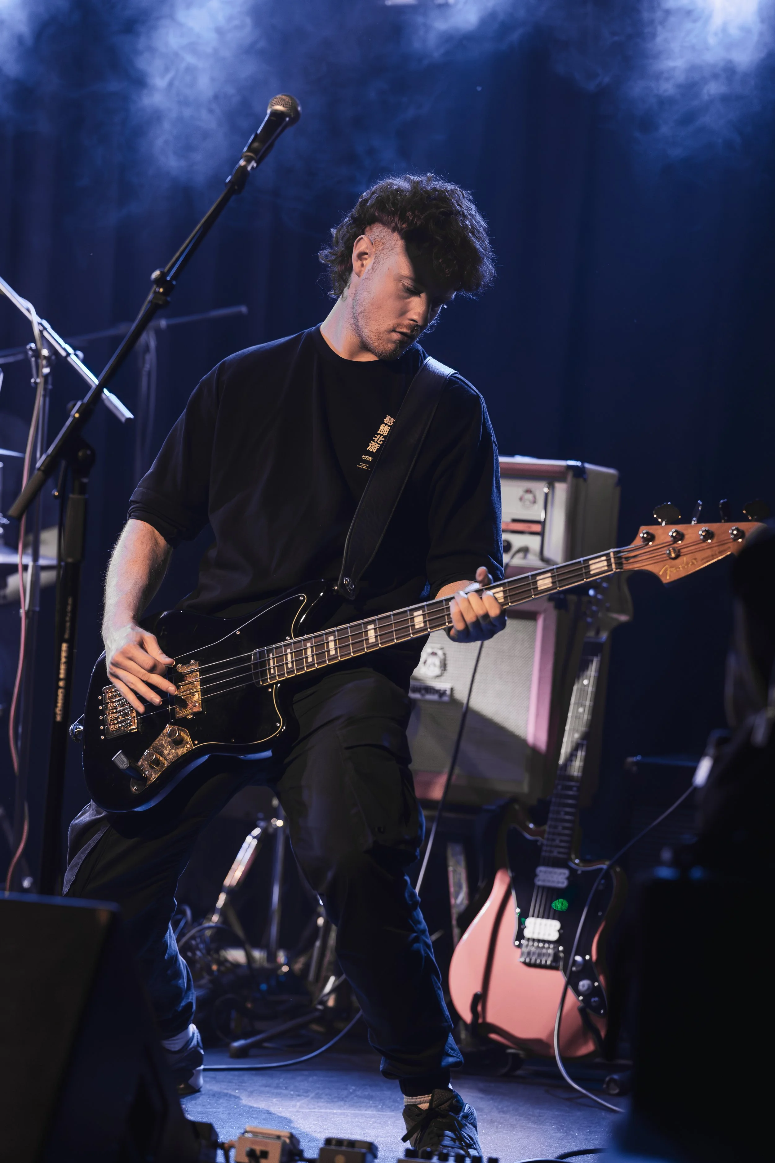 A young male musician playing a black electric bass guitar on stage, with music equipment and amplifiers behind him, under blue stage lighting.