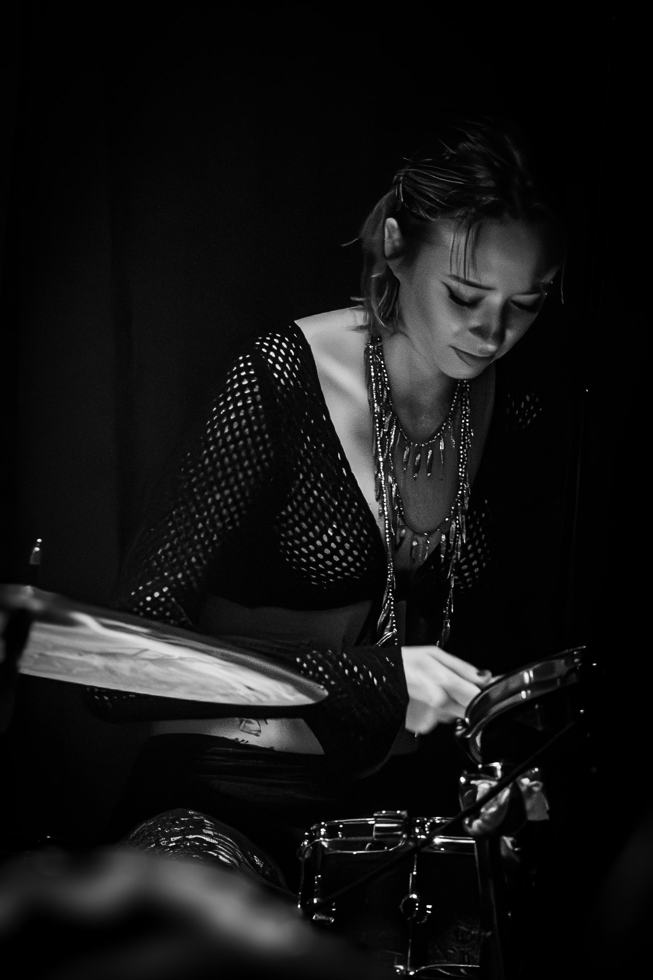 A woman with short hair in a black, fishnet top and layered necklaces, looking down and playing a drum set in a dark setting.