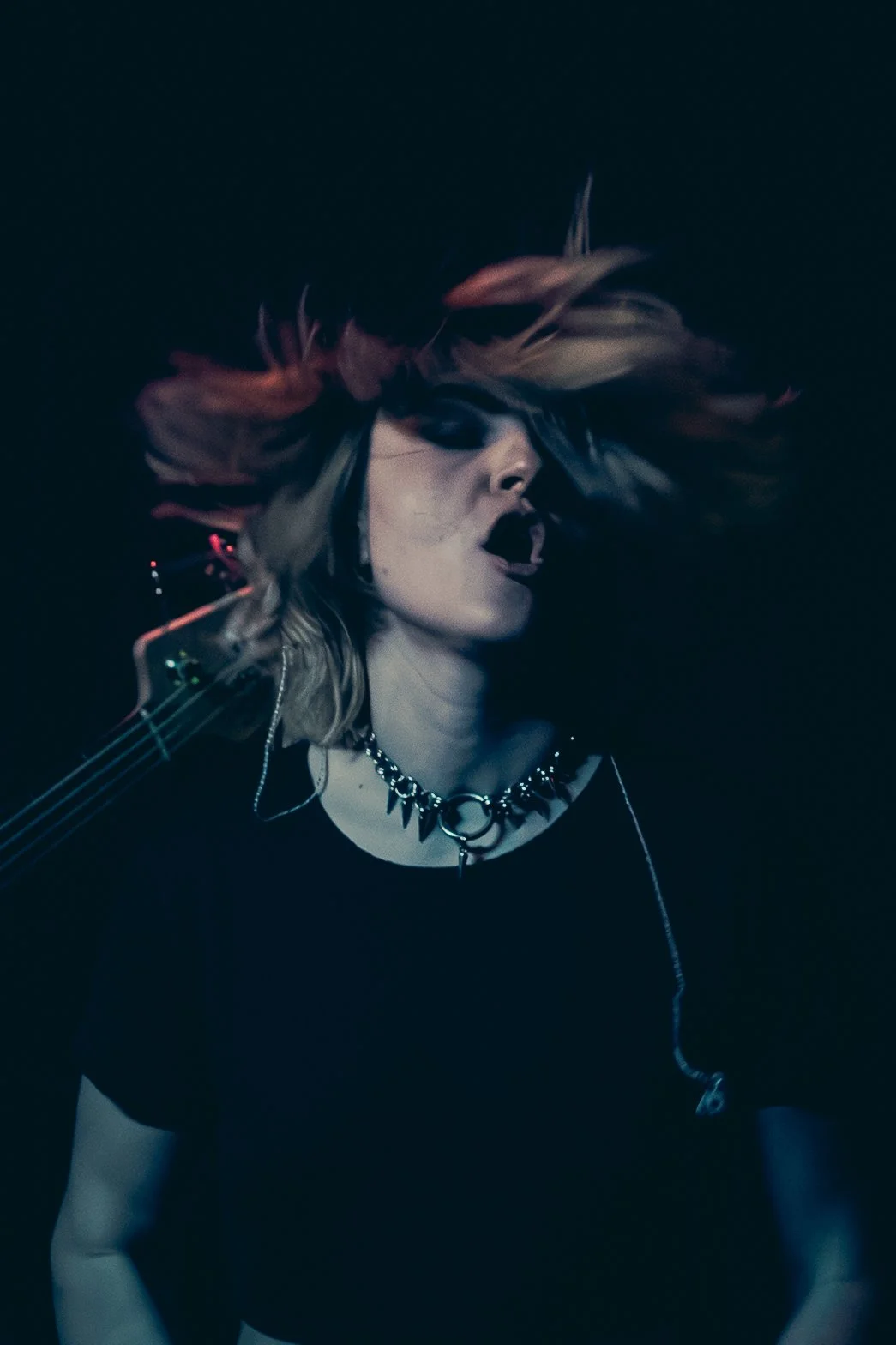 A woman with platinum blonde hair with pink tips is singing or performing, wearing dark makeup, a choker necklace, and a black shirt, with a guitar strap over her shoulder in a dark setting.