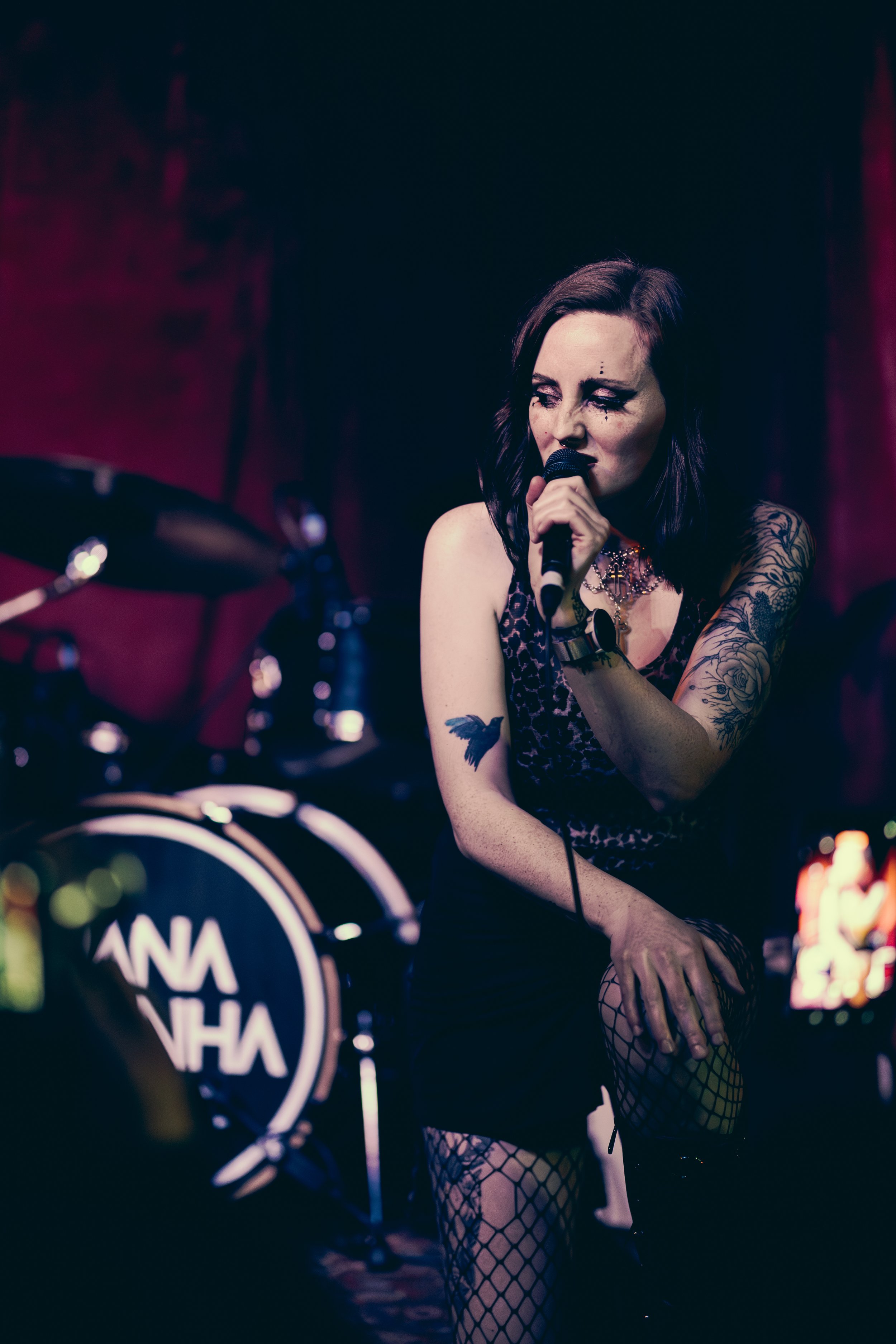 A woman with dark hair and tattoos passionately sings into a microphone on stage, with a drum set in the background.