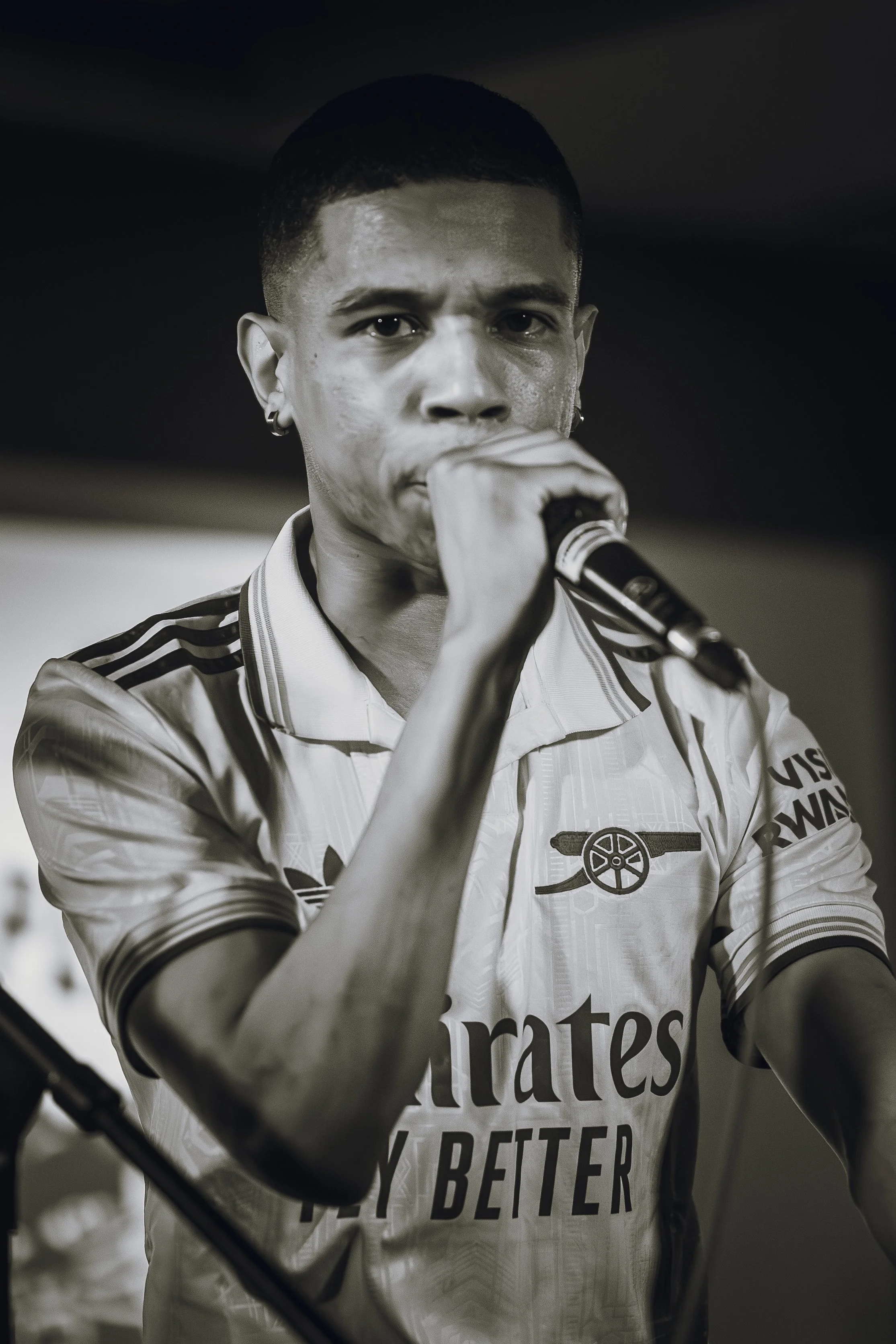 A person with a short haircut wearing earrings and a sports jersey, holding a microphone close to their mouth, looking directly at the camera.