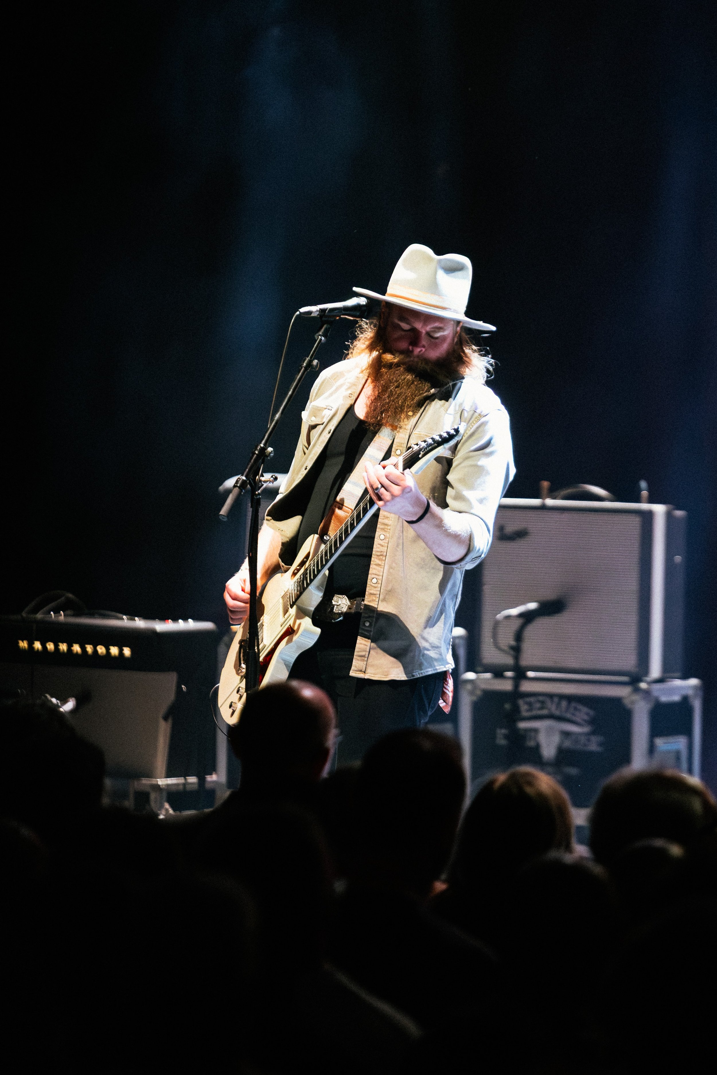 Musician wearing a cowboy hat playing an electric guitar on stage with amplifiers and equipment in the background, audience visible at the bottom.