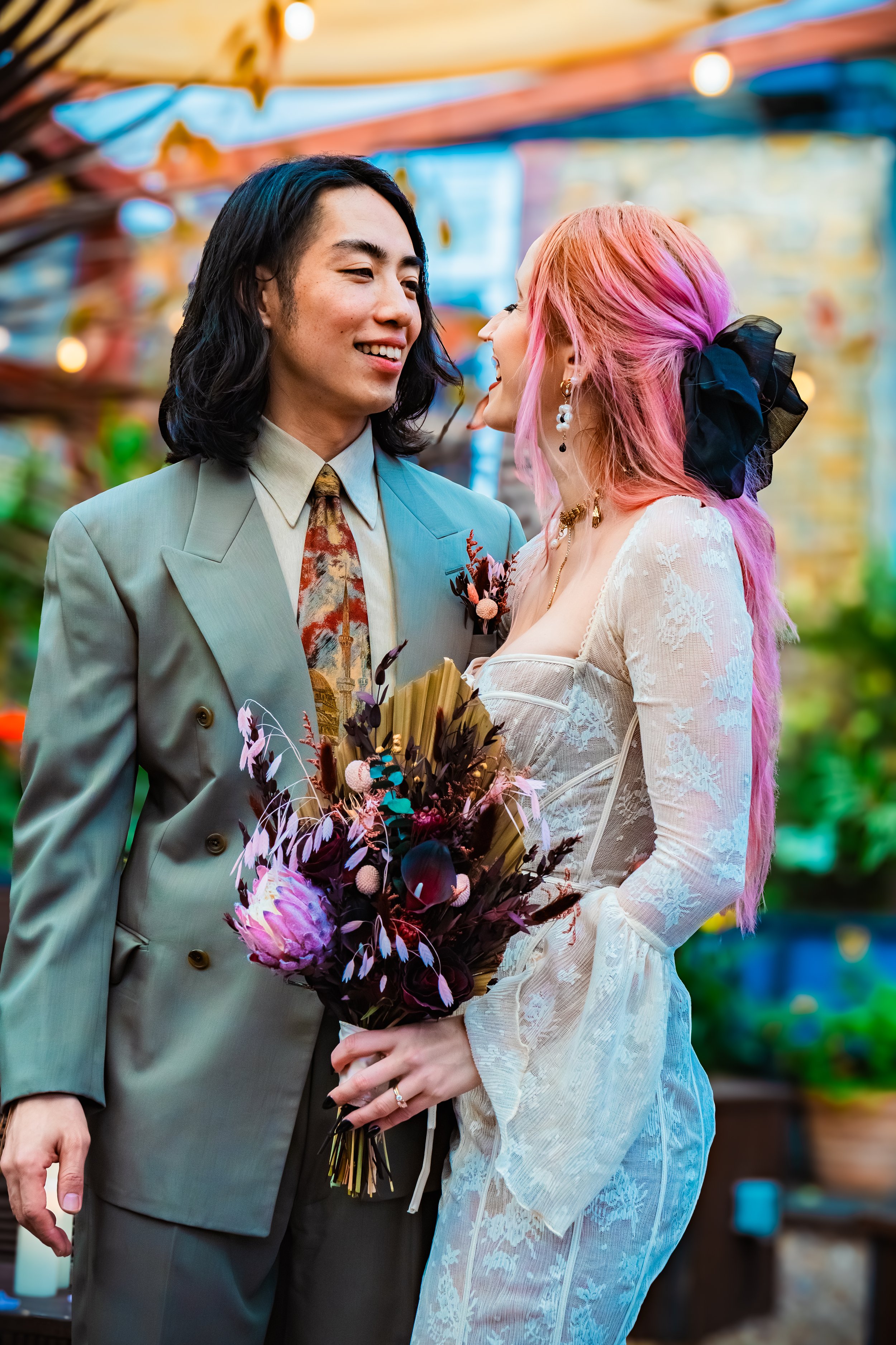 A couple dressed in wedding attire, standing close and smiling, with the woman holding a bouquet of purple and pink flowers, in an outdoor setting with colorful decorations.