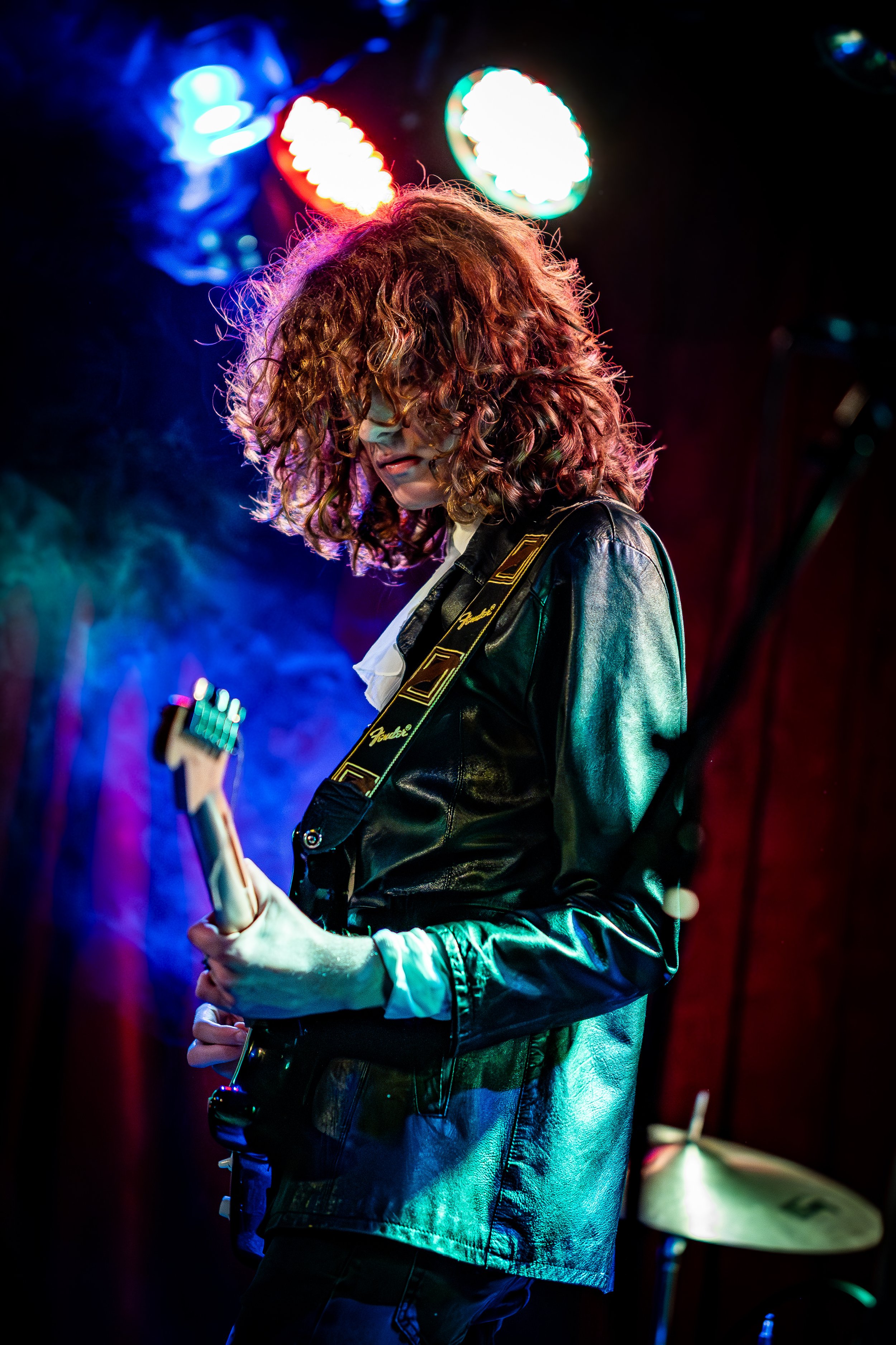 A woman with curly hair playing an electric guitar on stage, wearing a black leather jacket surrounded by colorful stage lights.