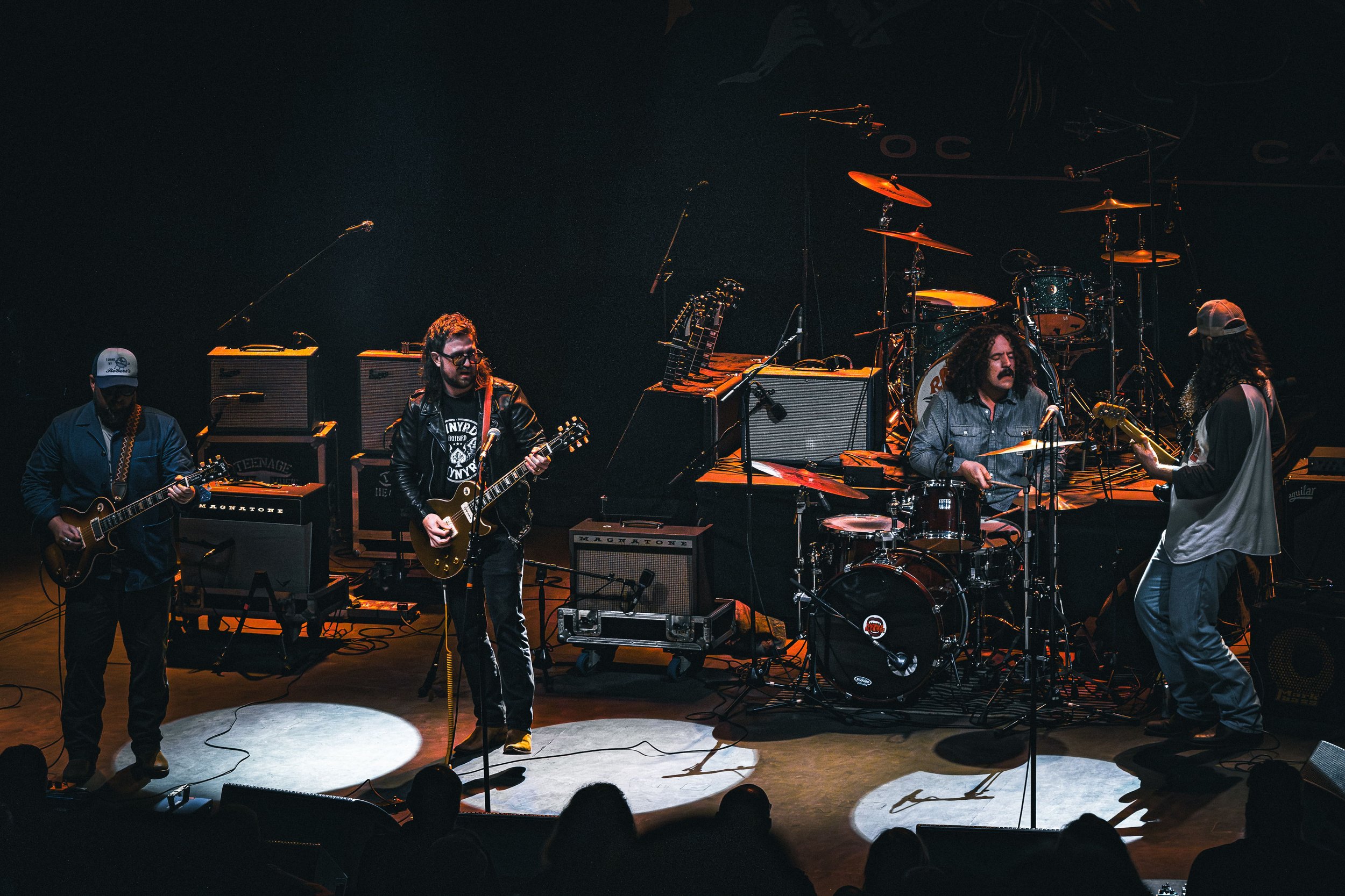 Four musicians perform on stage with guitars, drums, and amplifiers, with a dark background and stage lighting.