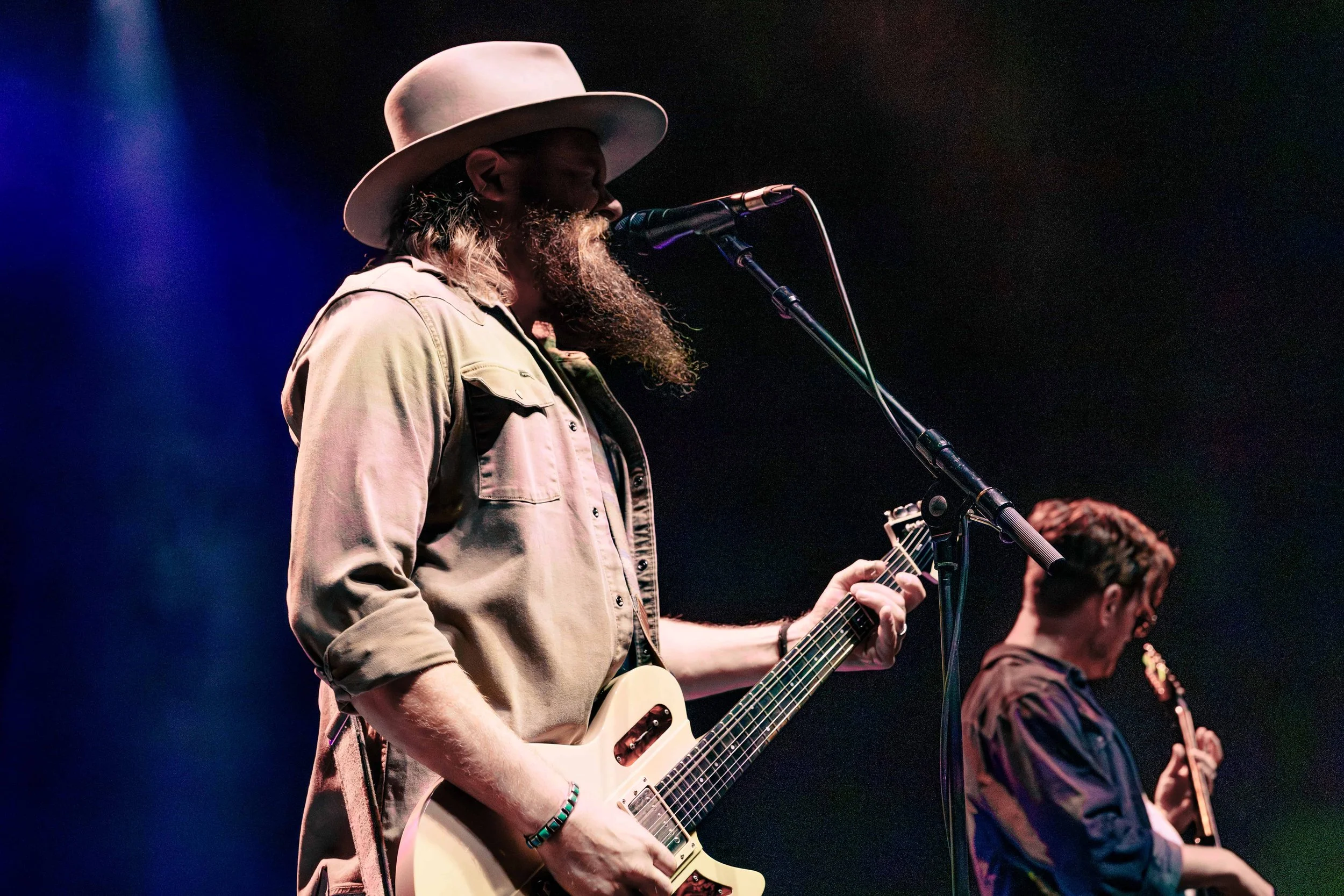 A musician with a beard and long hair wearing a wide-brimmed hat and beige jacket, playing an electric guitar and singing into a microphone on stage with colorful lighting.