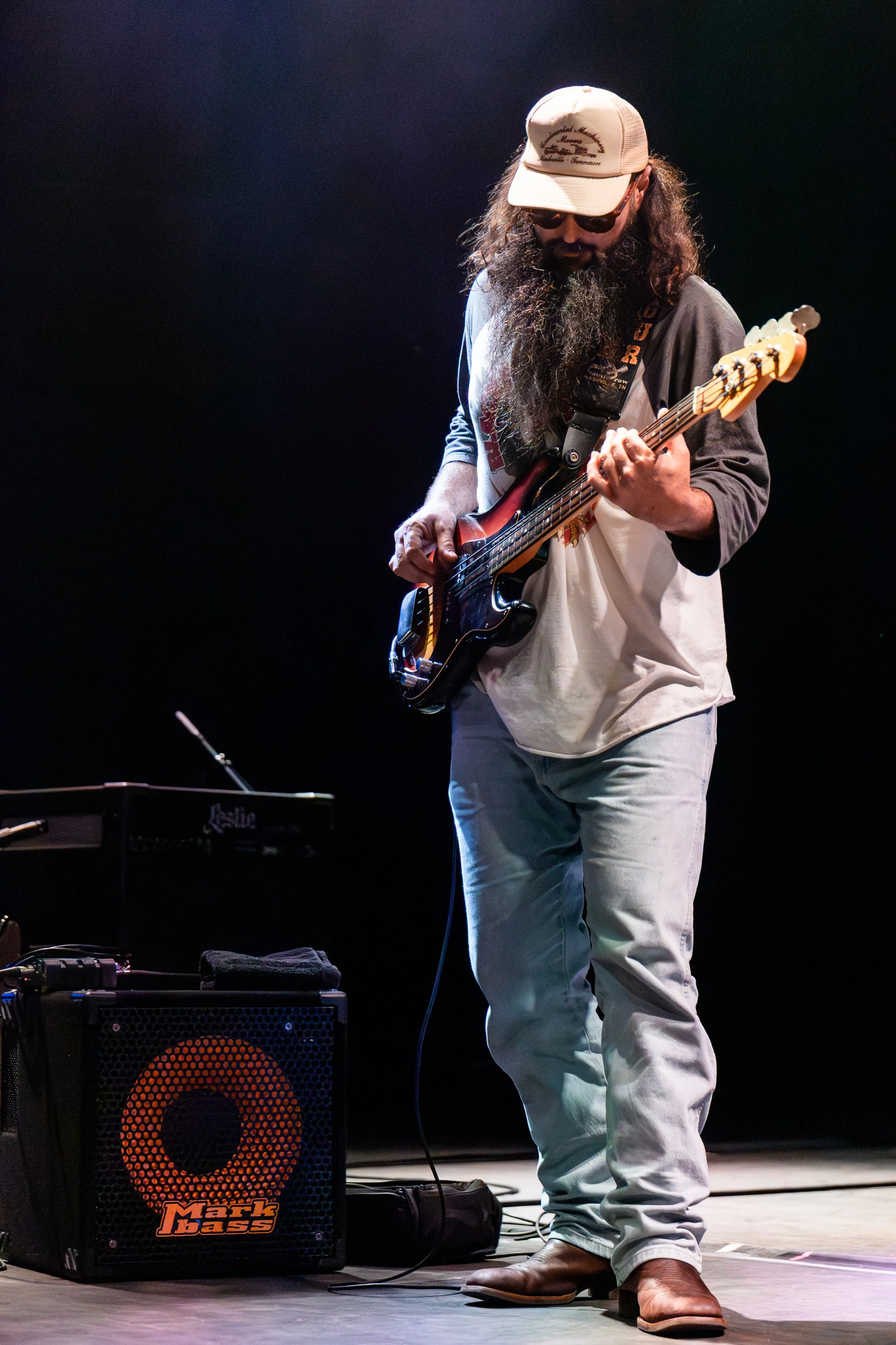 Man playing bass guitar on stage, wearing a beige baseball cap, sunglasses, a gray and white jacket, light-colored pants, and brown boots, with musical equipment around him.