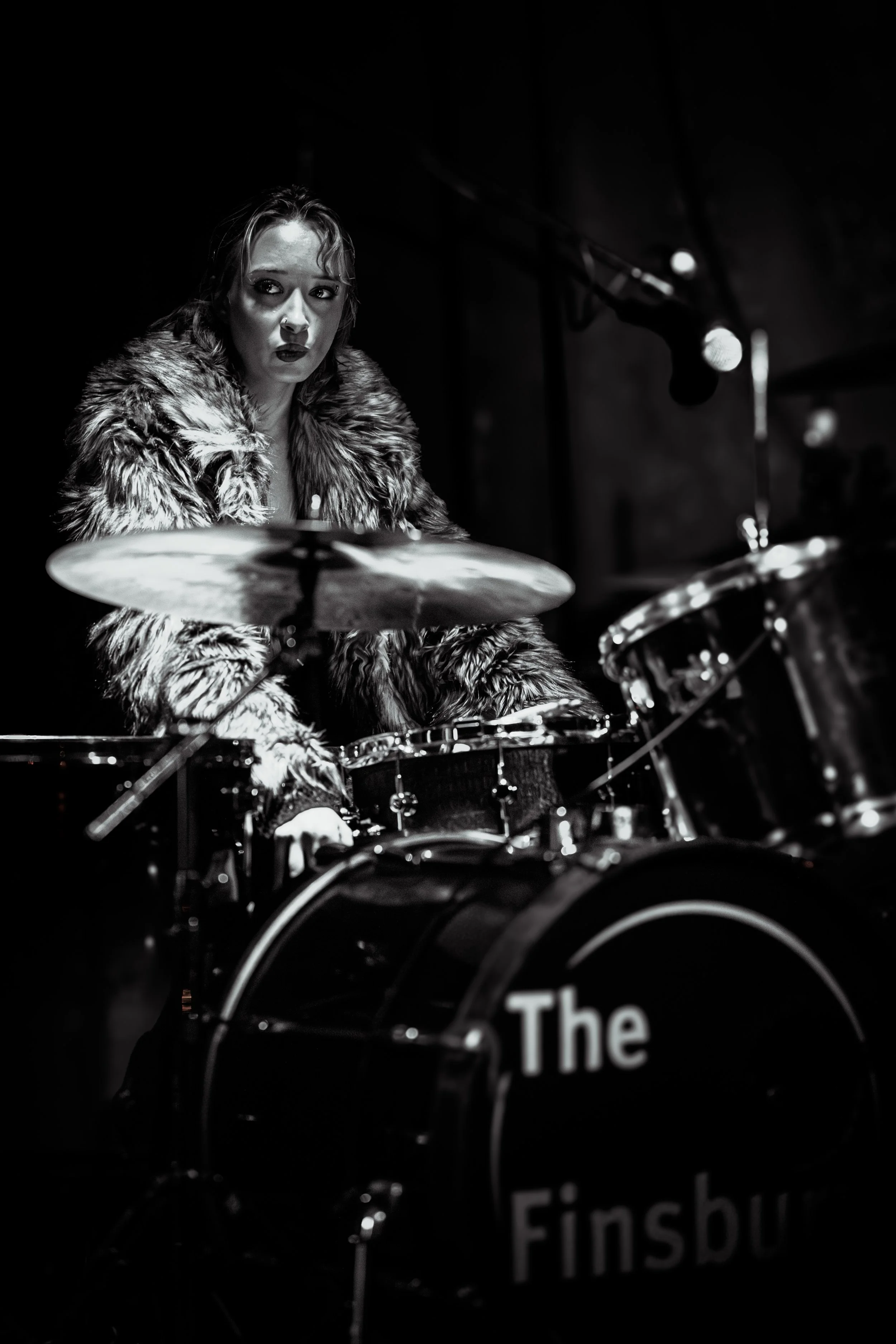 A black-and-white photo of a woman playing drums on stage, wearing a fur coat, with a focused expression, microphone overhead, and the drum set labeled "The Finsbu".