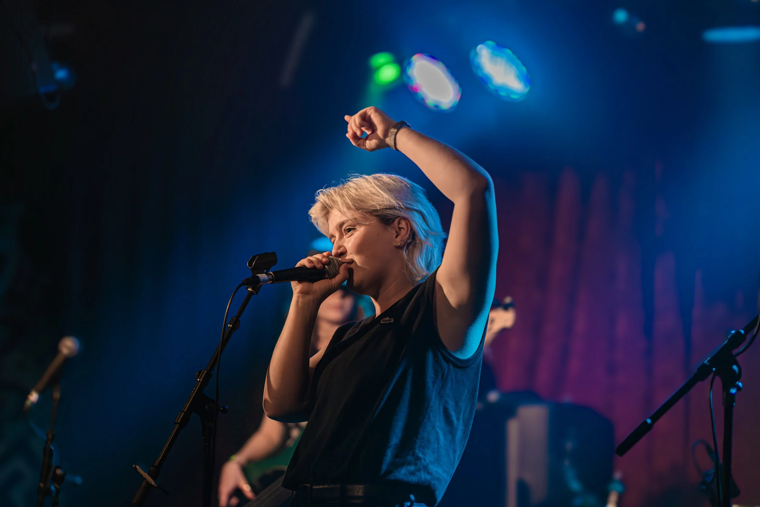 A female singer with short blonde hair performing on stage with a microphone, with her right arm raised, under colorful stage lights.