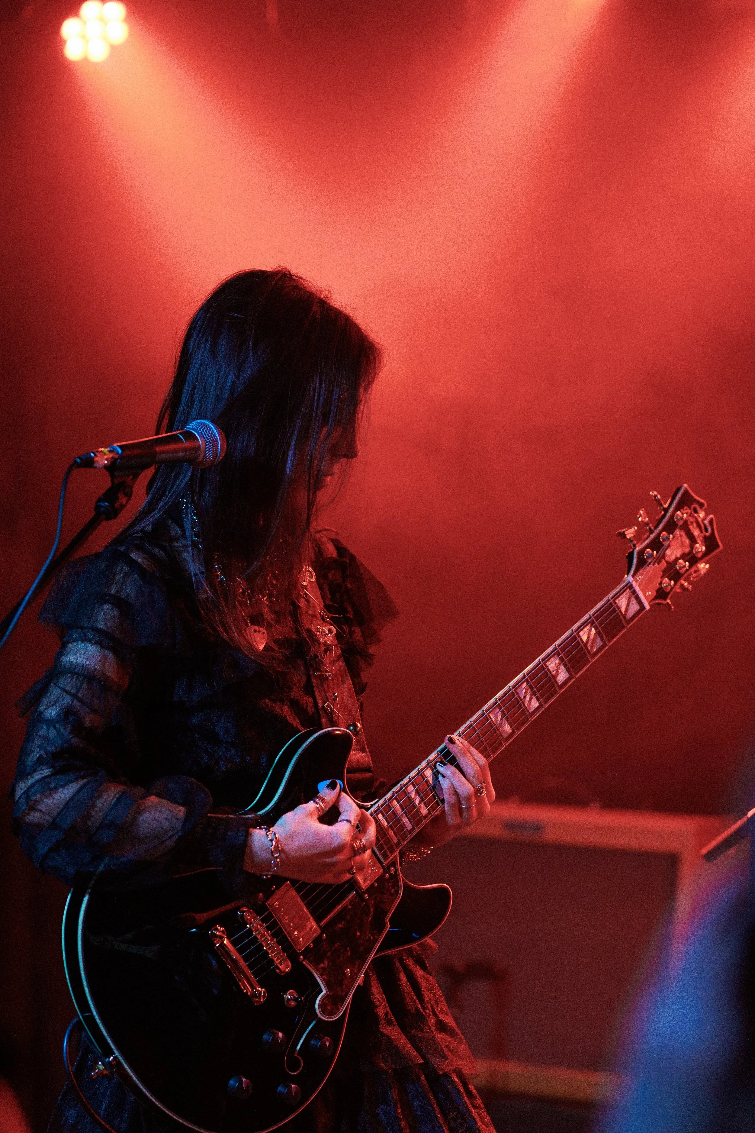 A woman with long dark hair playing an electric guitar on stage under red lighting with a microphone nearby.