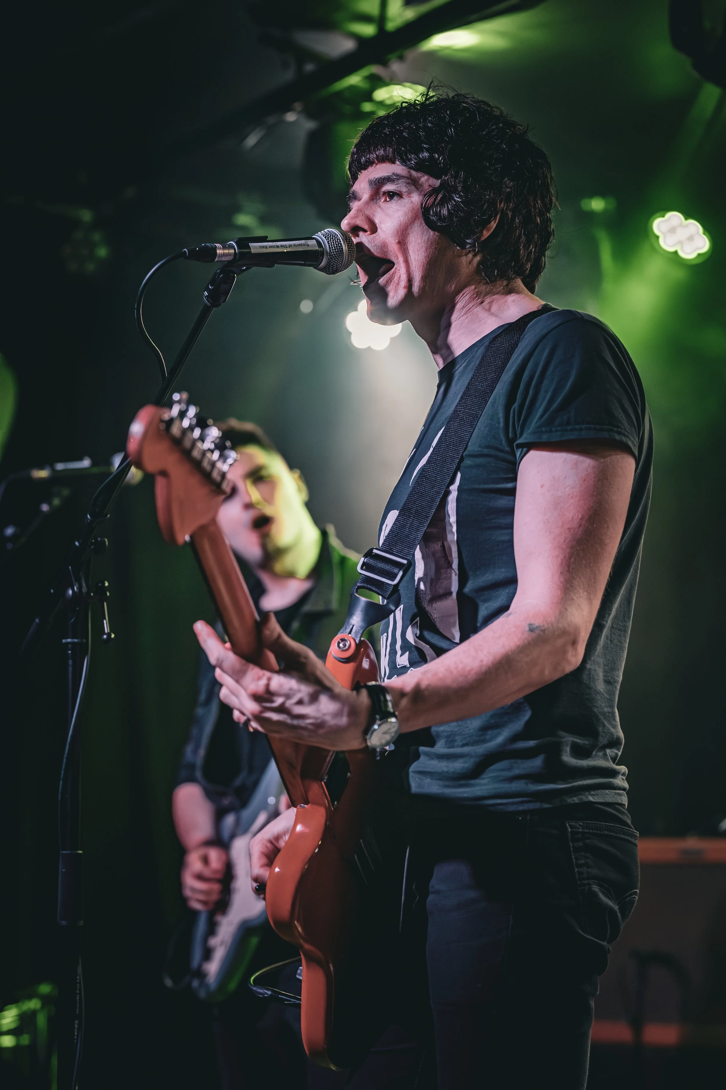 A musician singing into a microphone while playing an electric guitar on stage with colorful lighting.