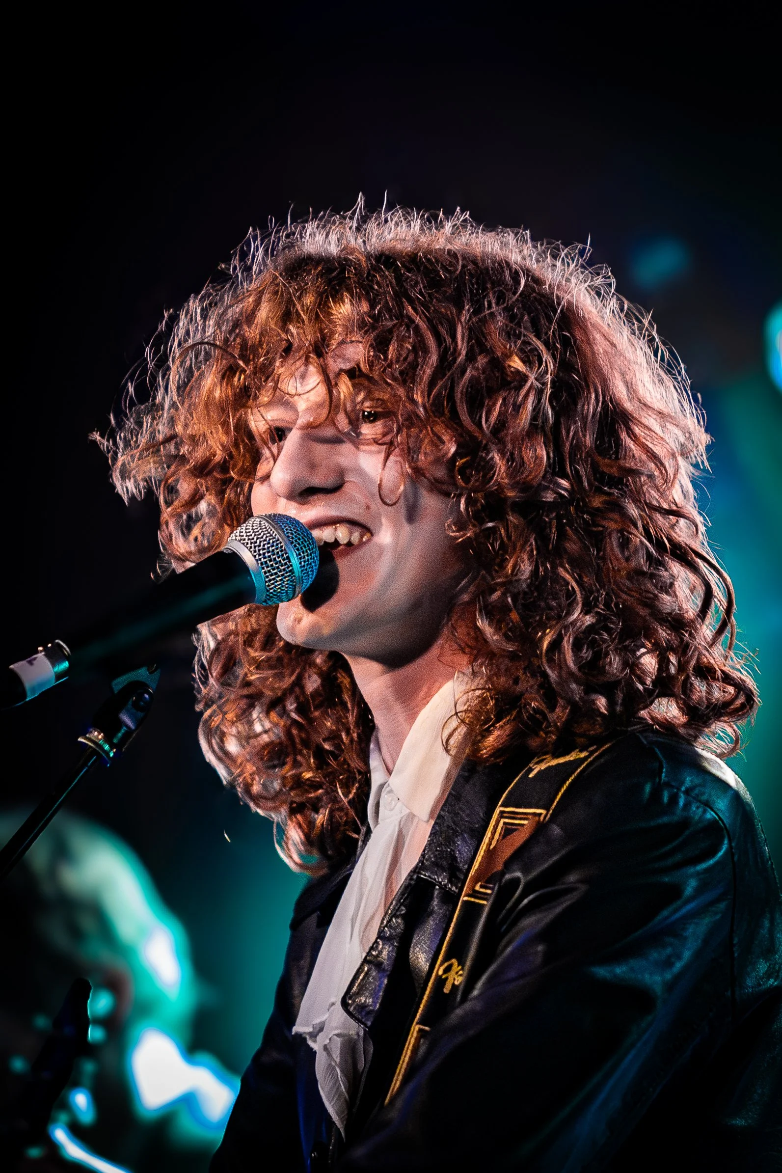 A musician with curly red hair singing into a microphone, wearing a leather jacket and white shirt, performing on stage.