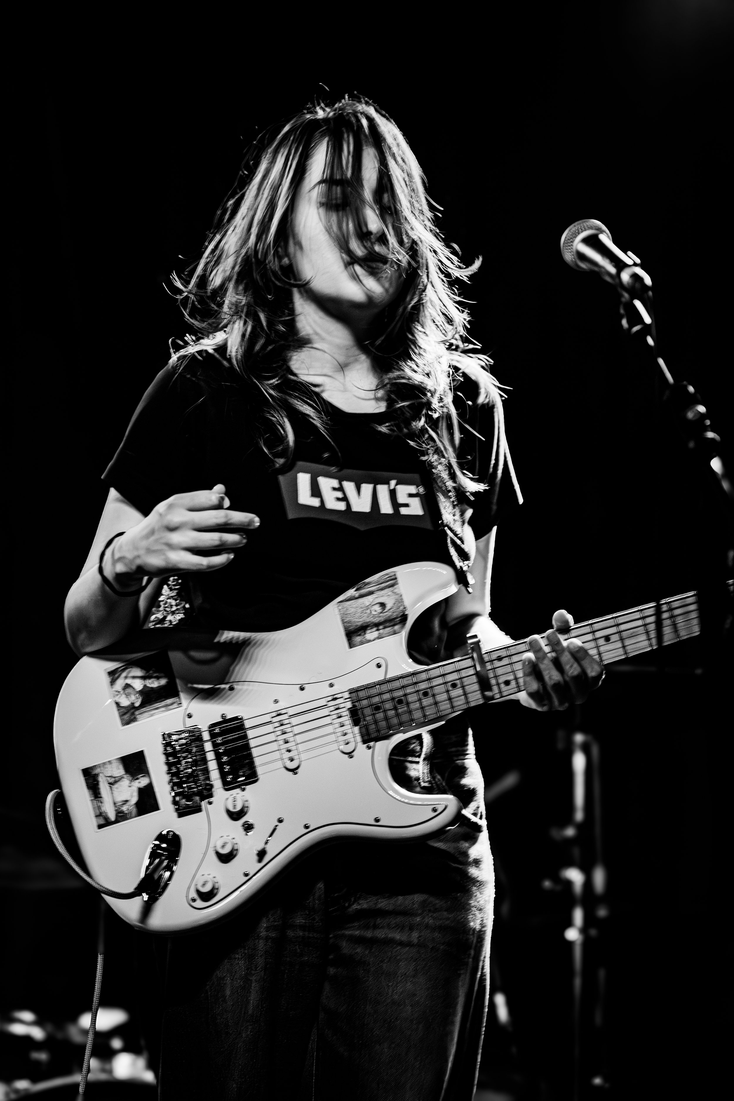 Black and white photo of a female musician playing an electric guitar on stage, wearing a Levi's t-shirt with her hair partially obscuring her face.