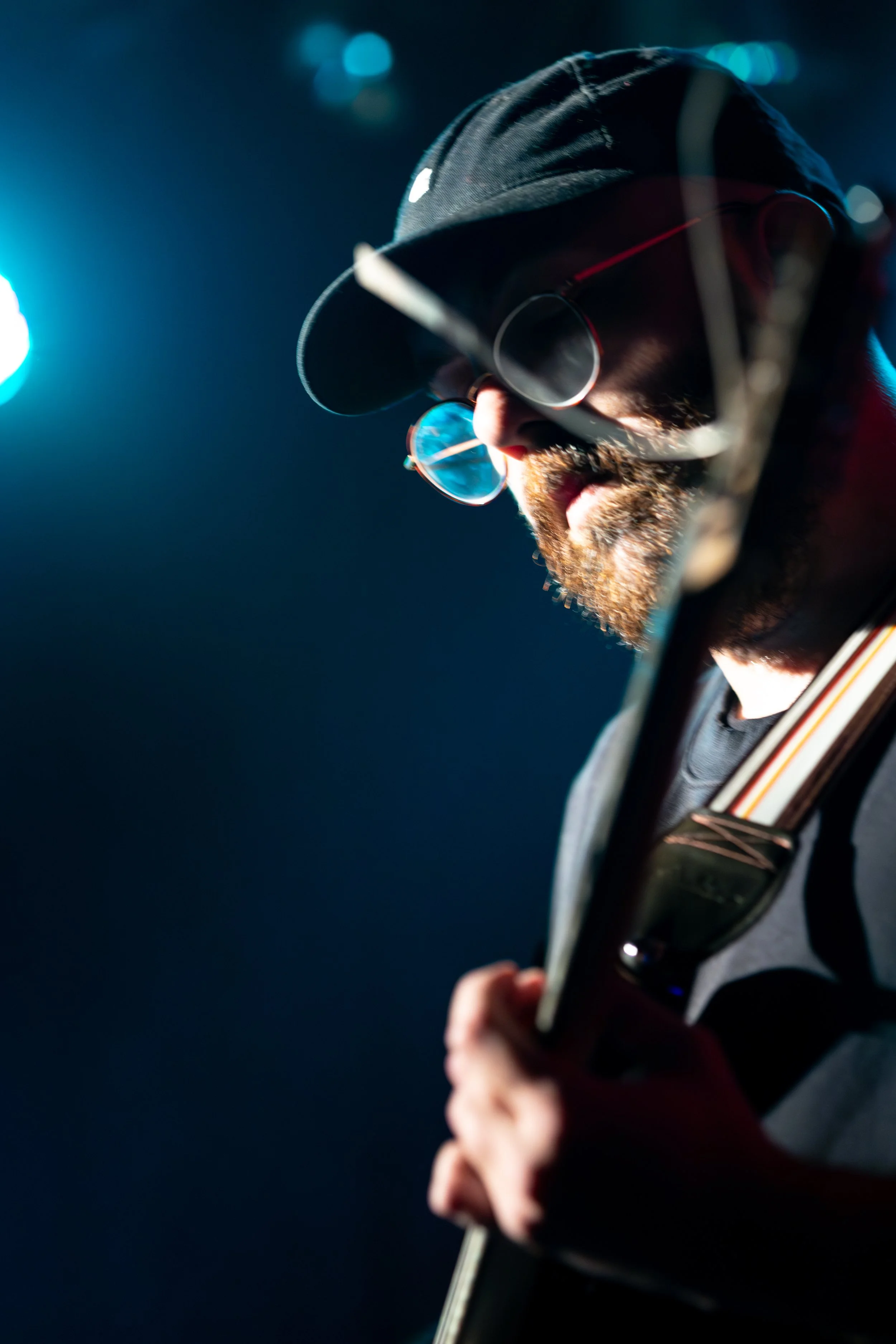 A man wearing a black cap, sunglasses, and a beard playing an electric guitar on stage with dramatic blue lighting.