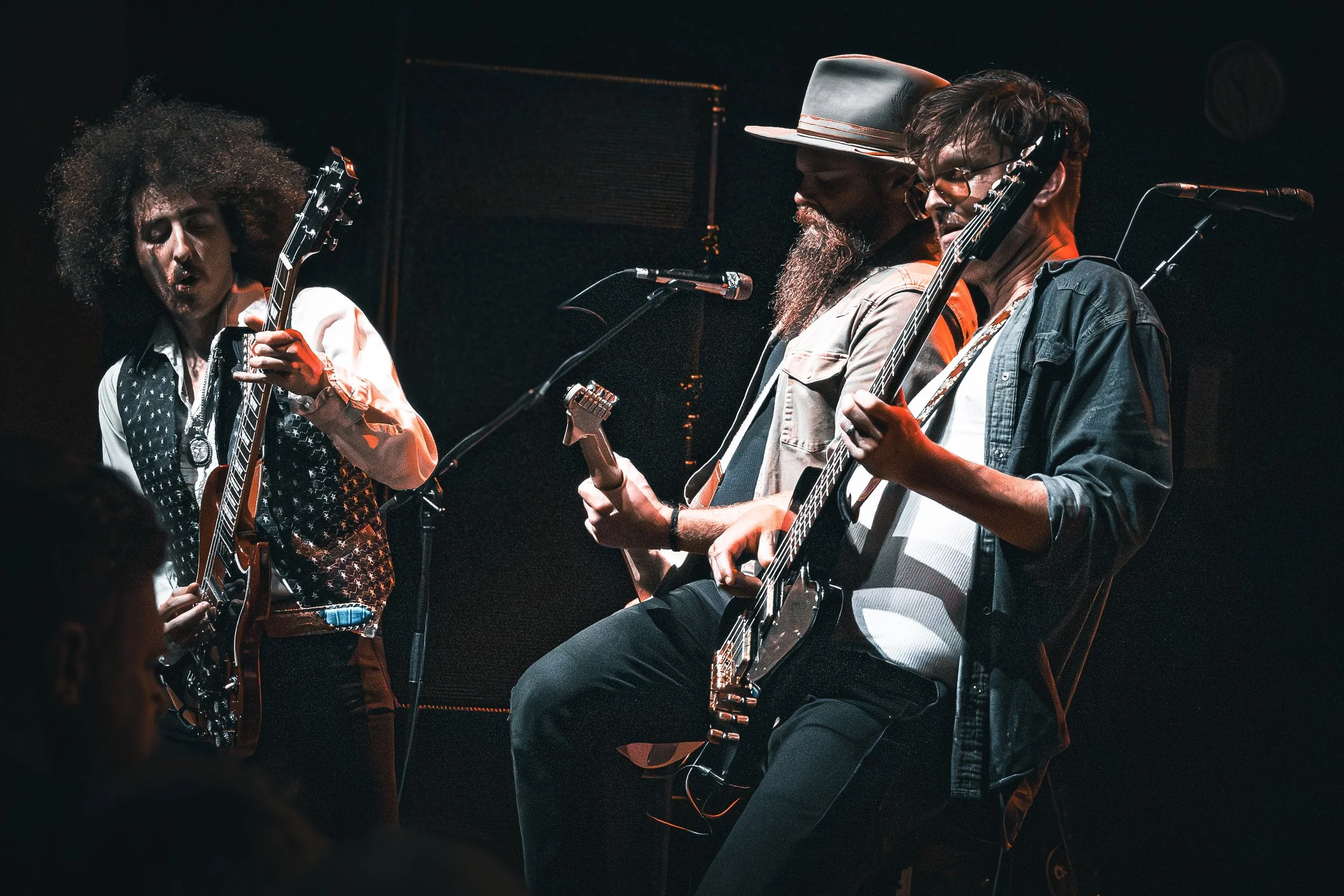 Three musicians performing on stage, playing guitars and singing into microphones, with dark background lighting.