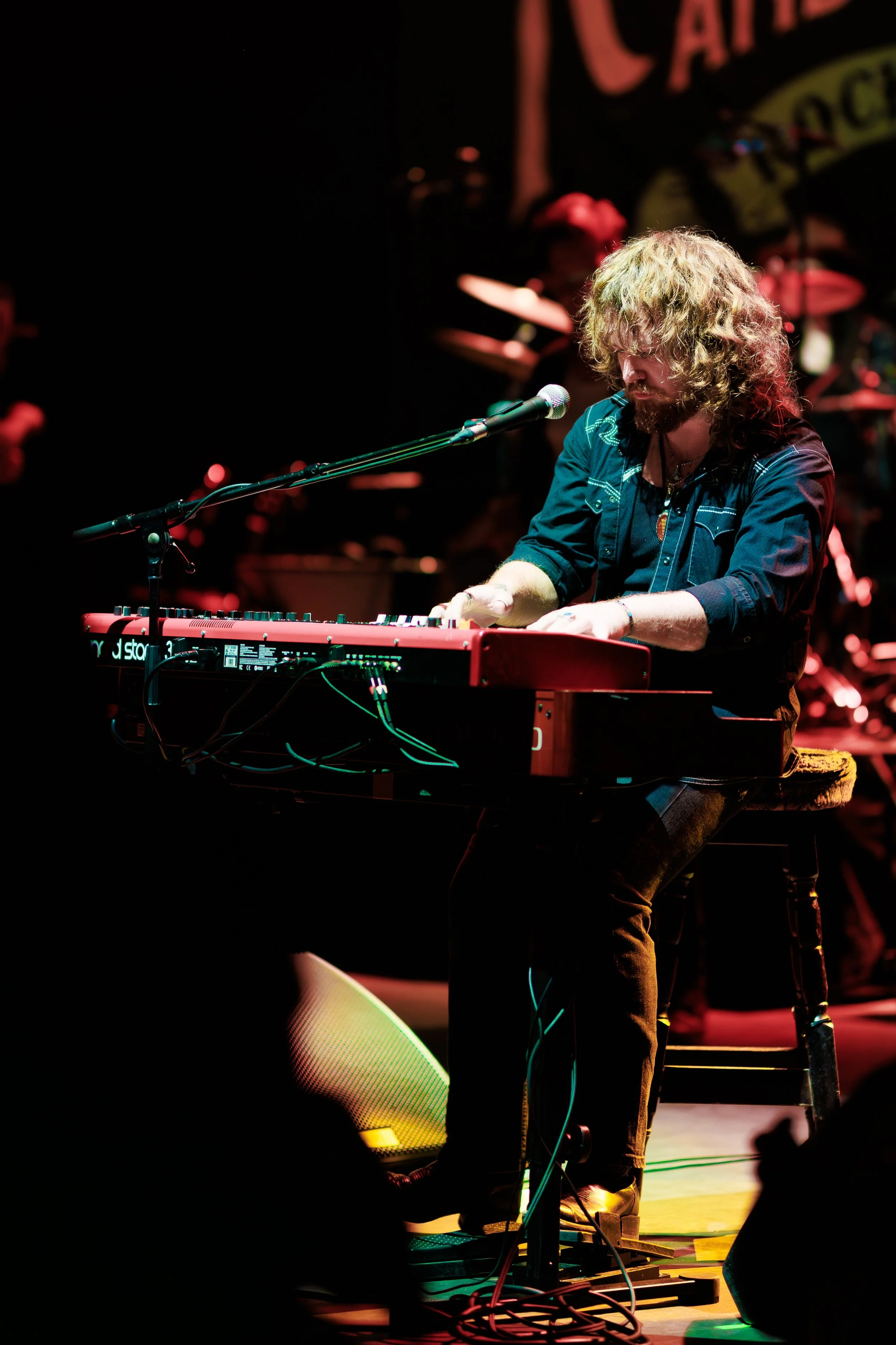 A musician with curly hair and a beard playing a keyboard on stage, with a microphone in front of him, and a drummer partially visible in the background, surrounded by stage lights.