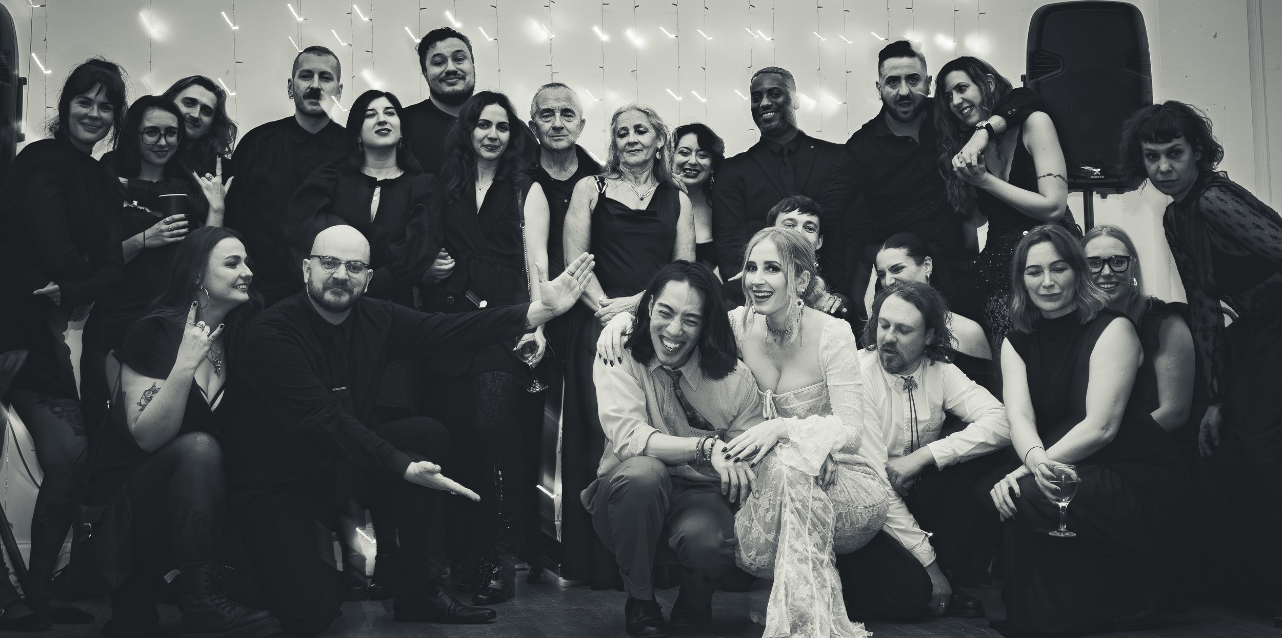 A large group of people, diverse in age and style, gathered together at a celebration or event, smiling and posing for a group photo in front of a decorated backdrop.