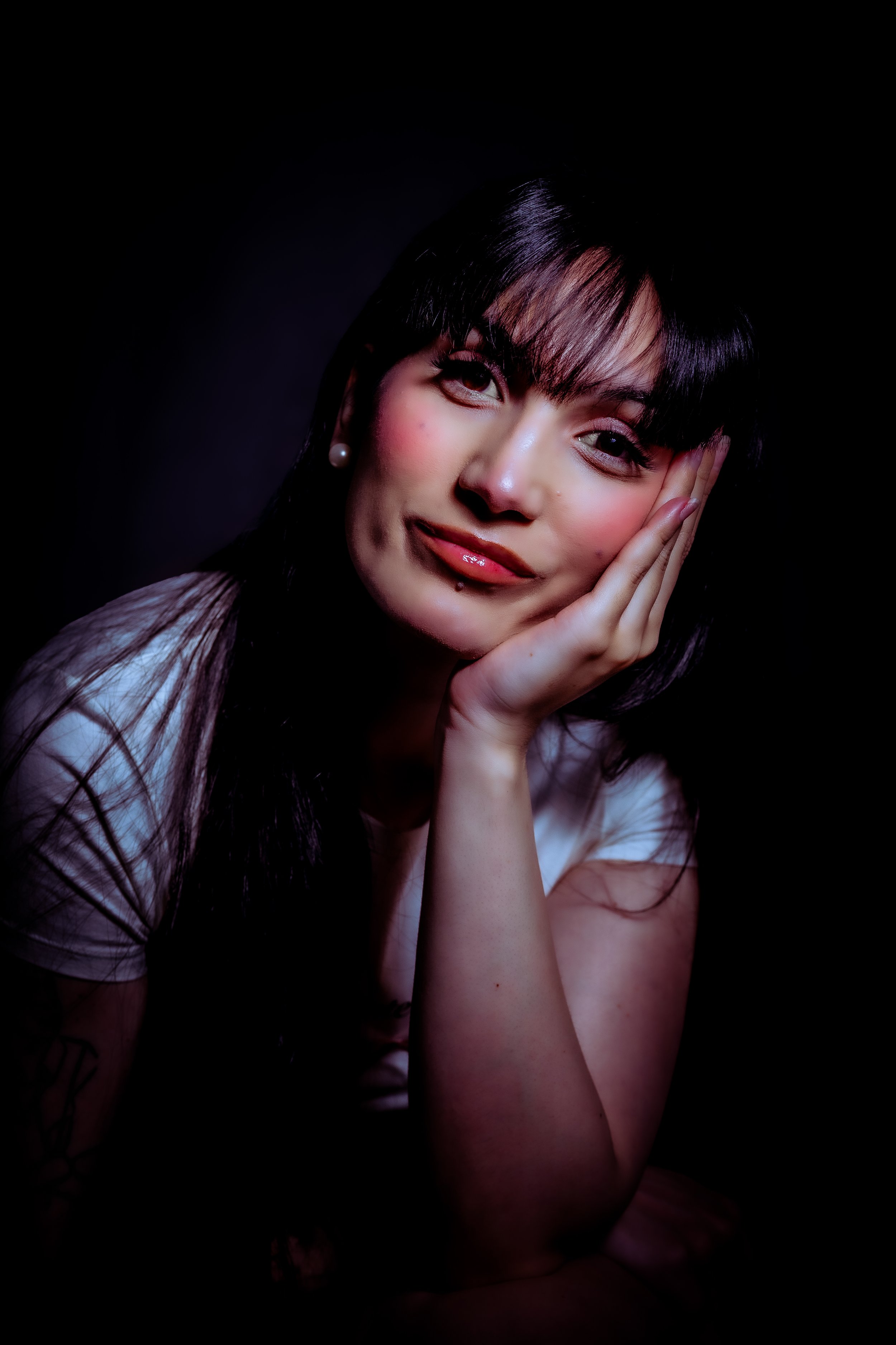 Portrait of a woman with dark hair, wearing pearl earrings and a white shirt, resting her face on her hand against a dark background.