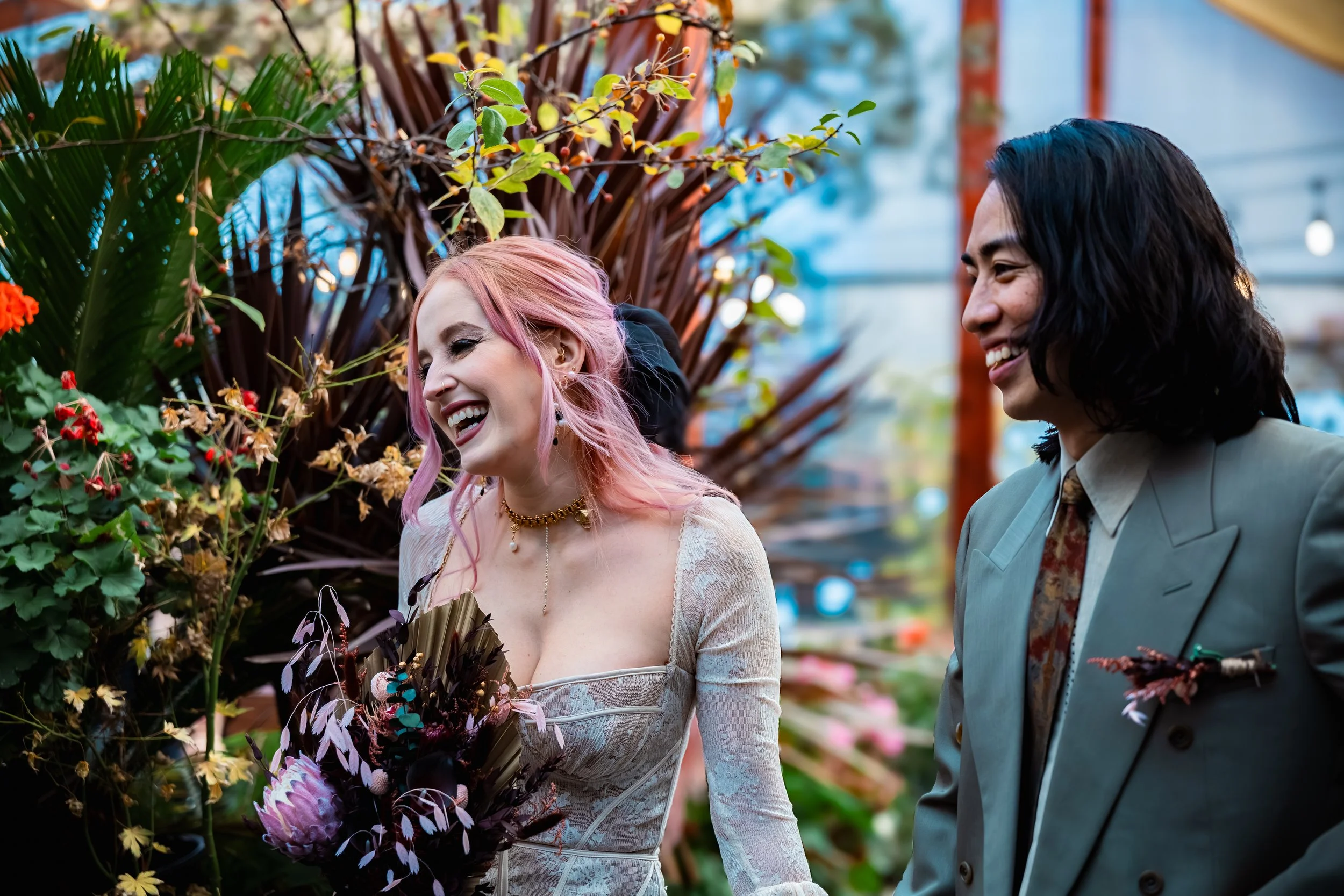 Two people smiling and laughing while standing near plants and flowers in a greenhouse or garden.