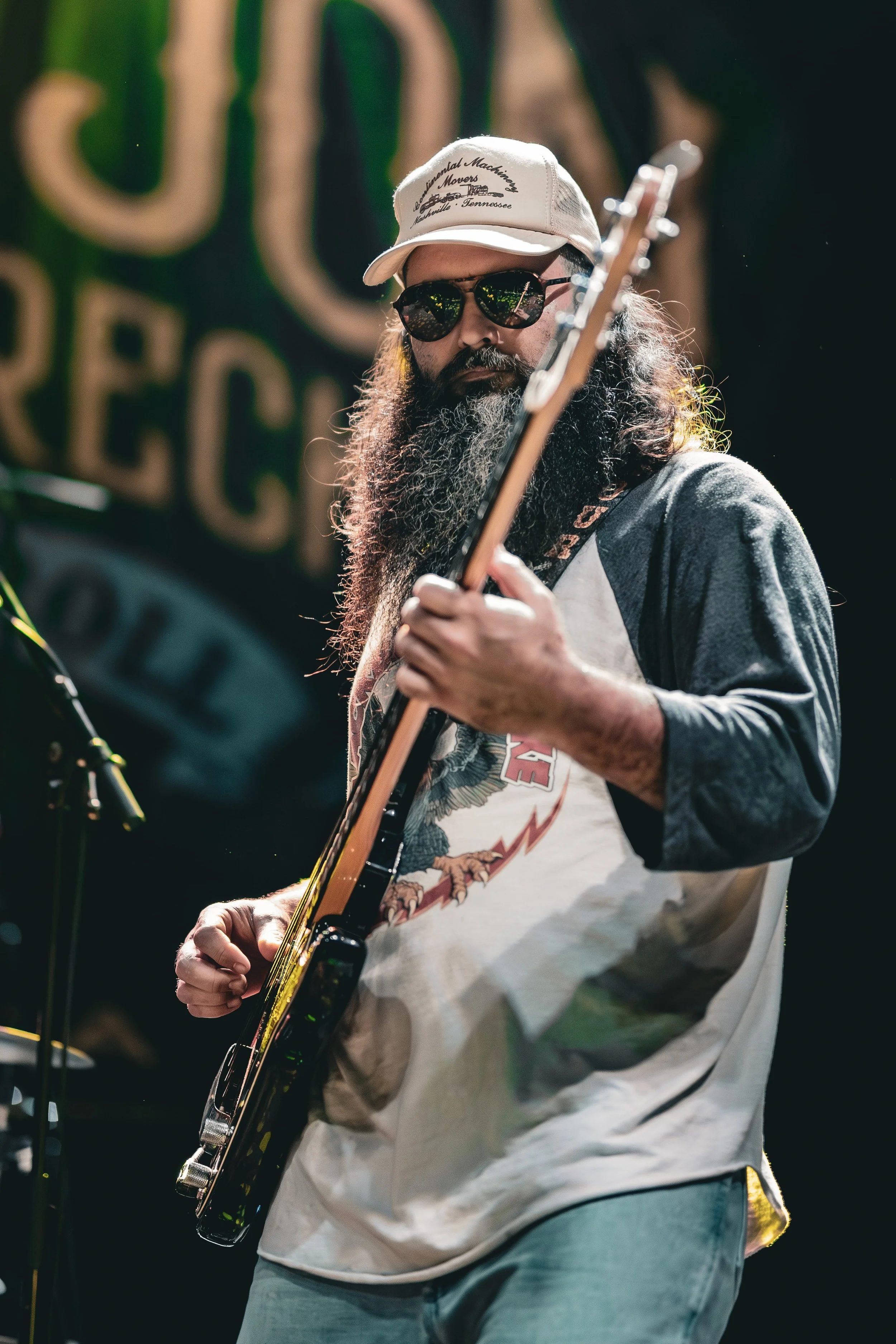 A man with long hair and a thick beard, wearing sunglasses, a baseball cap, a graphic T-shirt, and a gray sweatshirt, plays an electric guitar on stage with a dark background.