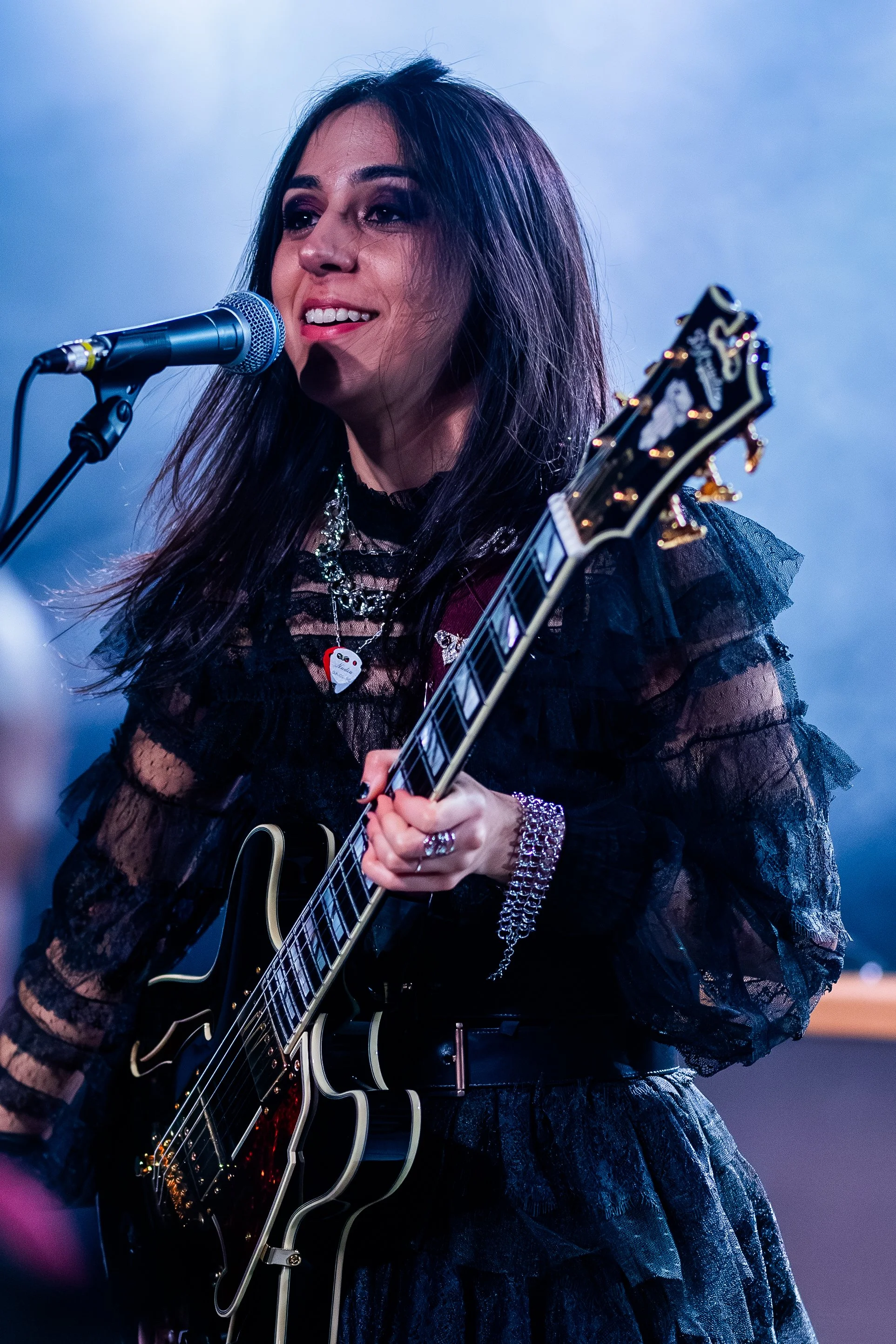 A woman with long dark hair and tattoos is singing into a microphone while playing a black electric guitar on stage. She is dressed in dark, lace clothing and wearing various jewelry.