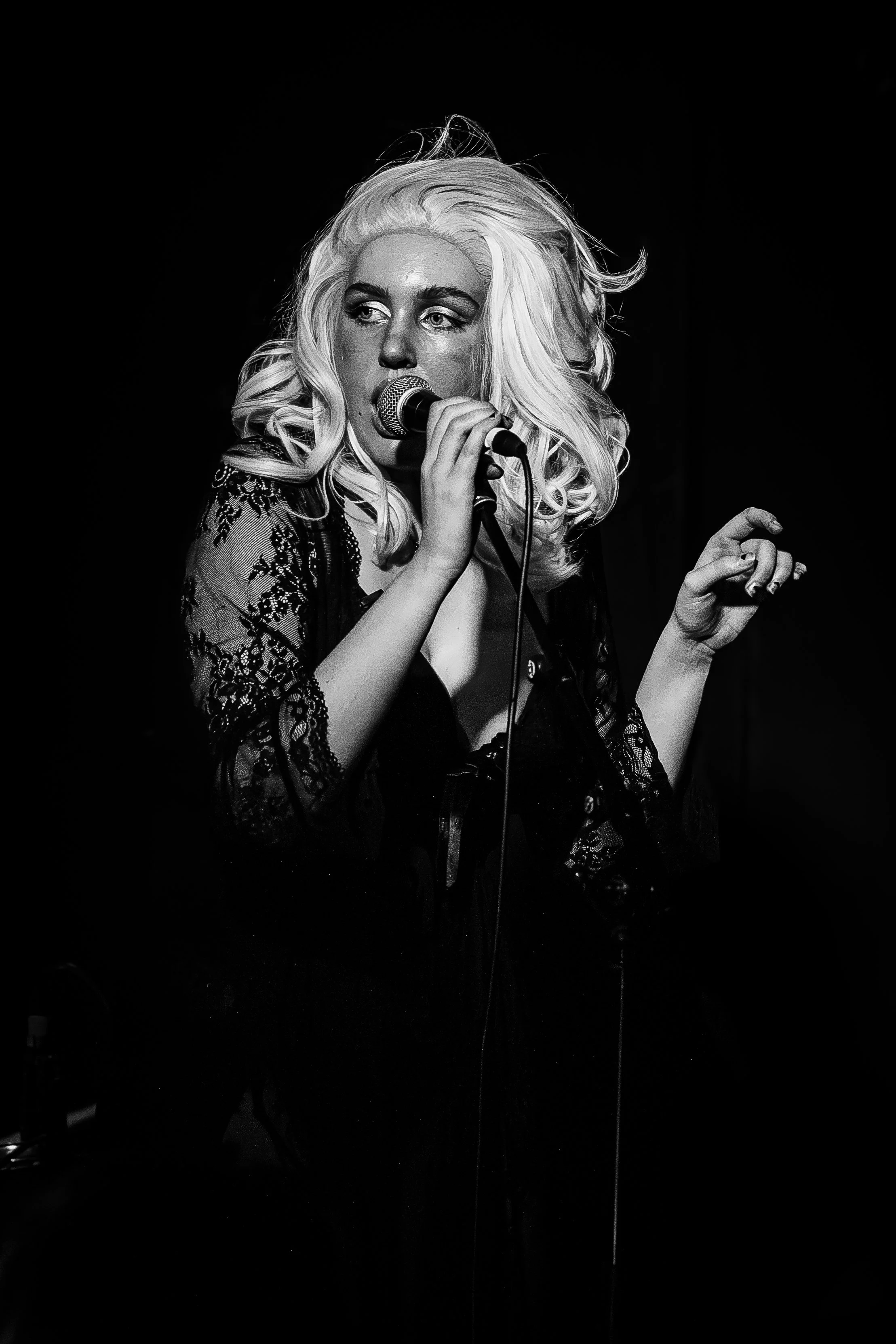 Black and white photo of a woman singing into a microphone on stage, with her eyes looking away, wearing a lace outfit and curly blonde hair.