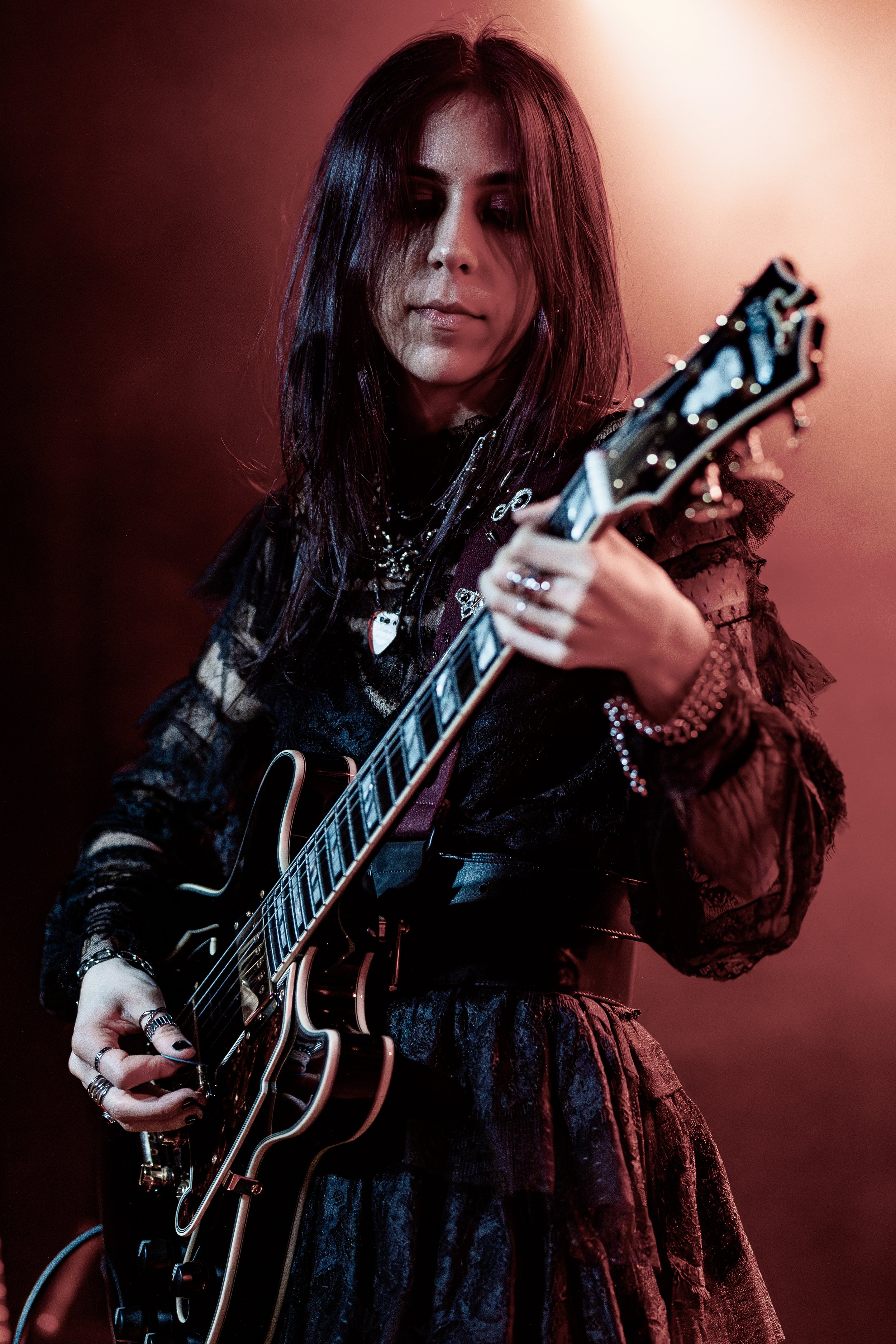 A woman with dark hair playing an electric guitar on stage, wearing a black lace outfit, surrounded by red stage lighting.