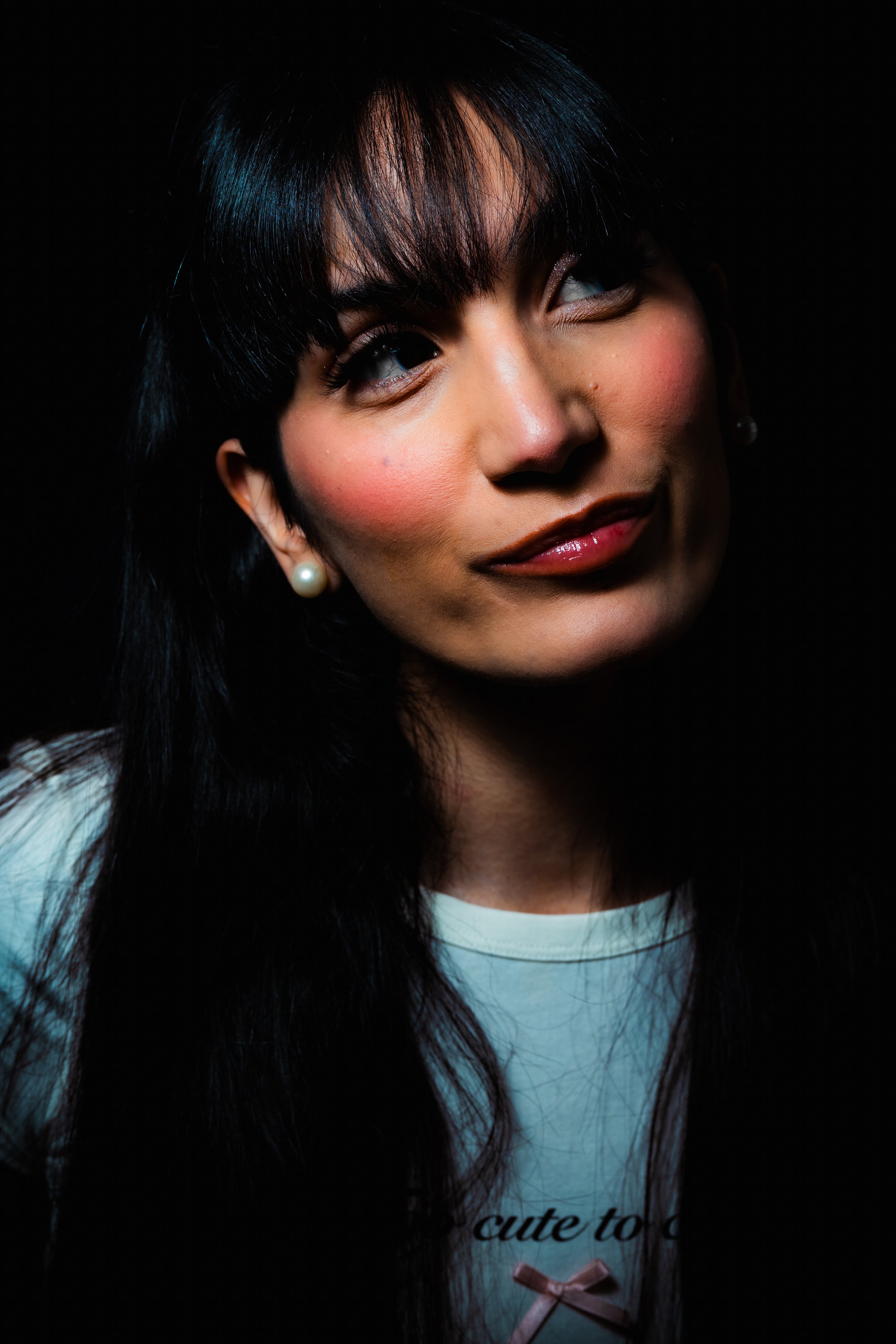 Close-up portrait of a woman with black hair, wearing pearl earrings, pink lipstick, and a white T-shirt with a small pink bow and the text 'cute to' visible.