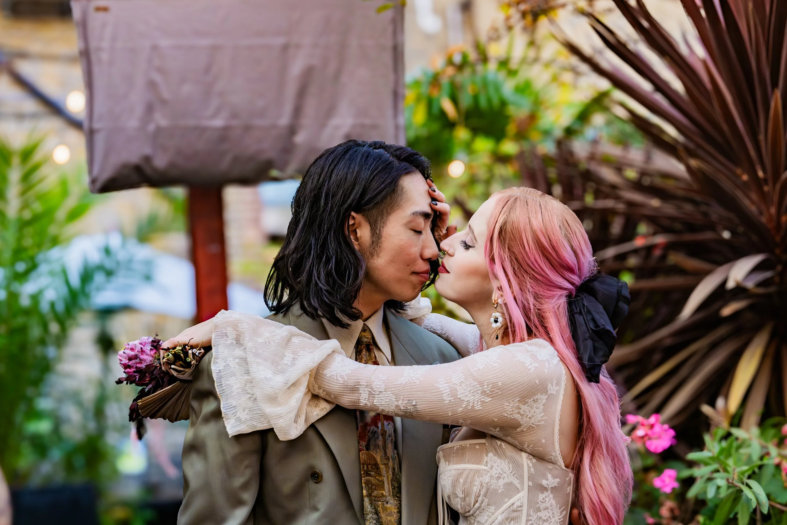 A couple, a man with long black hair and a woman with long pink hair, embraces and leans in for a kiss in a garden setting with greenery and flowers.