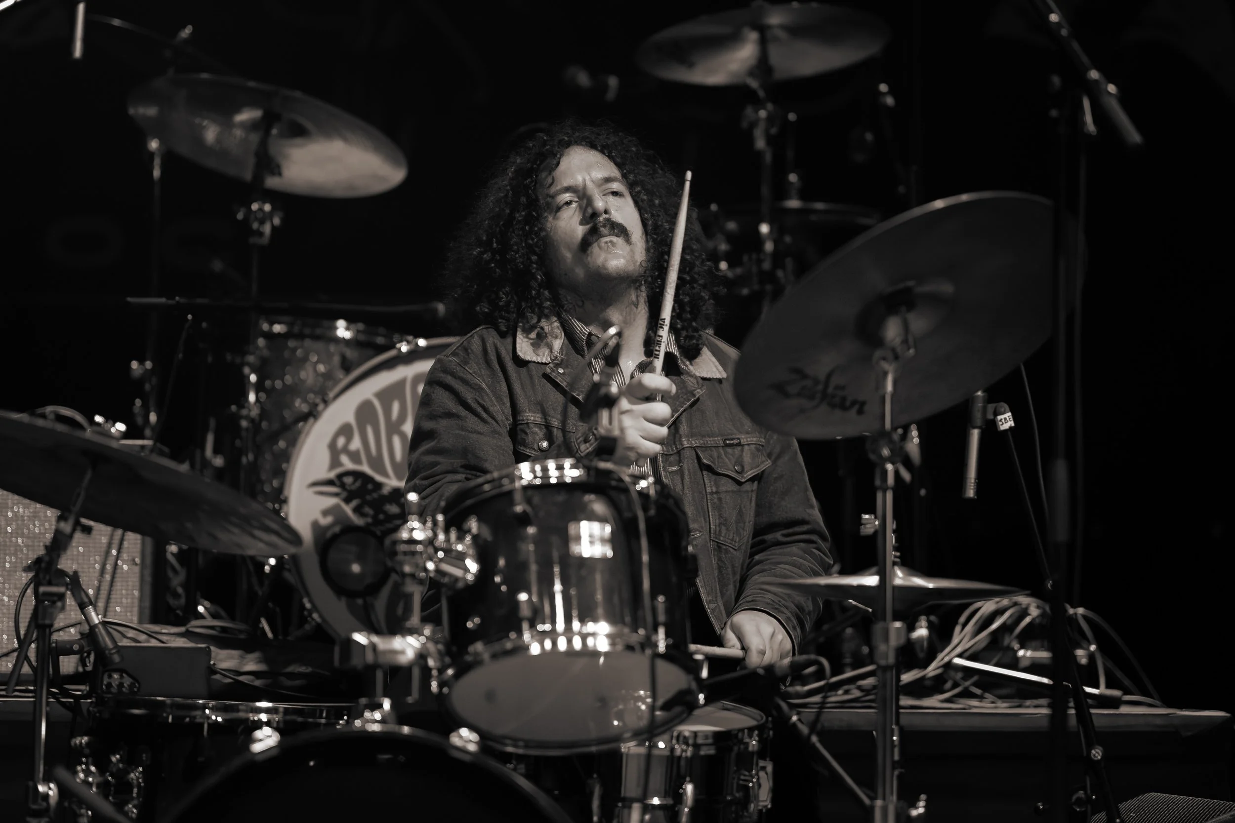 A man with long curly hair and a mustache playing a drum set on stage in black and white.