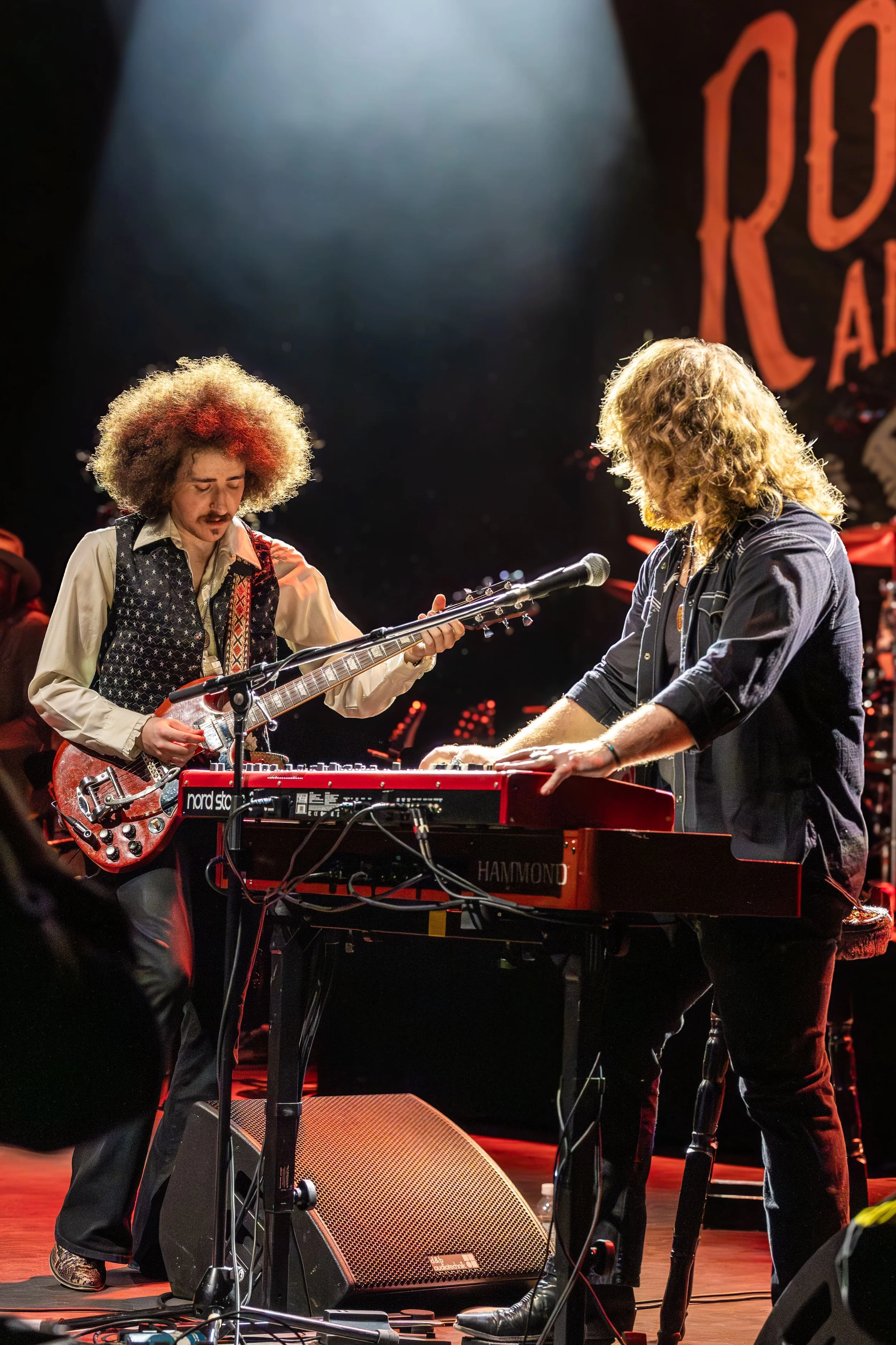 Two musicians perform on stage, one playing an electric guitar and the other playing a keyboard. They are focused on their instruments, and stage lighting highlights their figures.