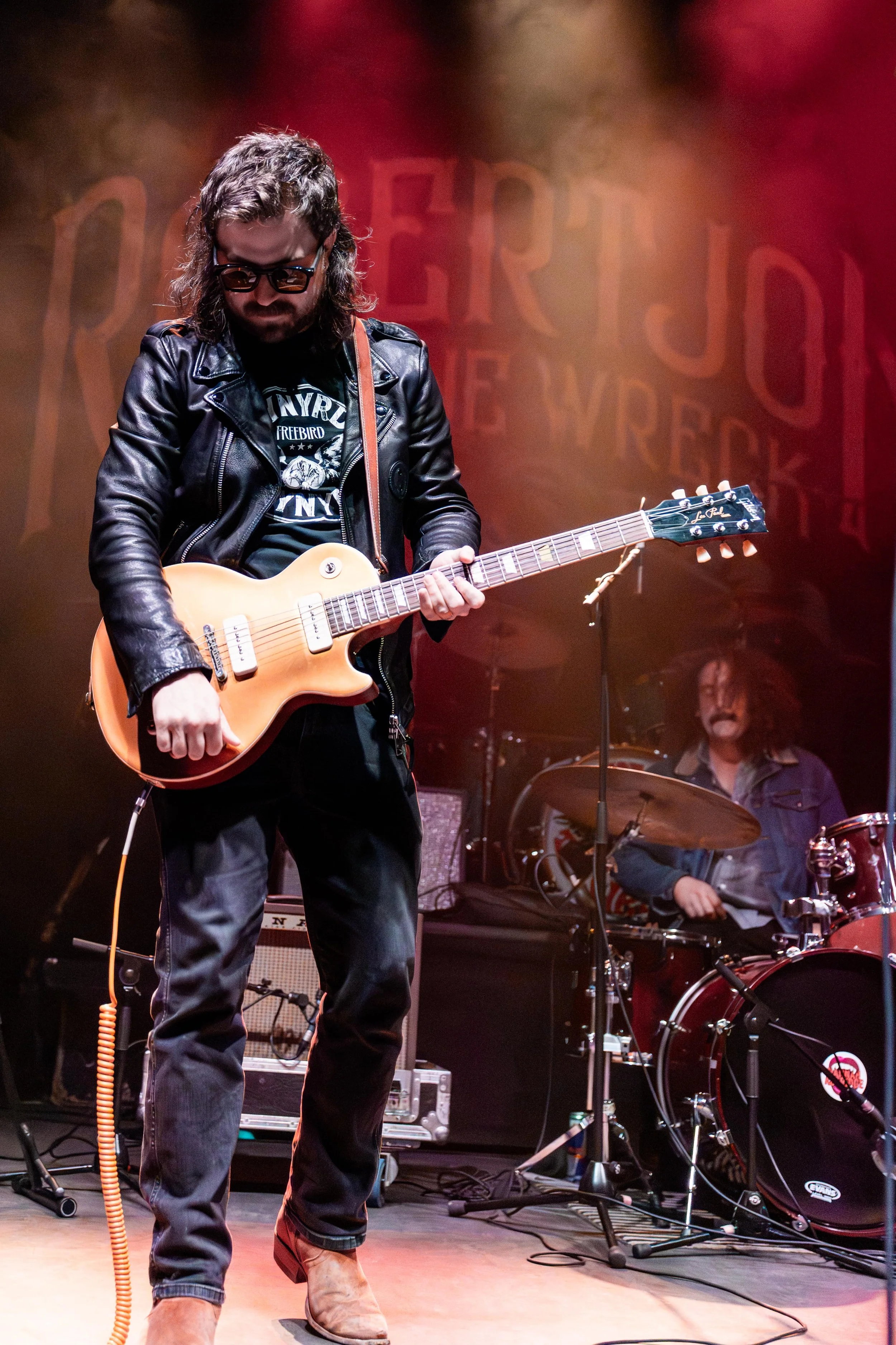 A musician playing an electric guitar on stage with a drummer in the background and stage equipment around.