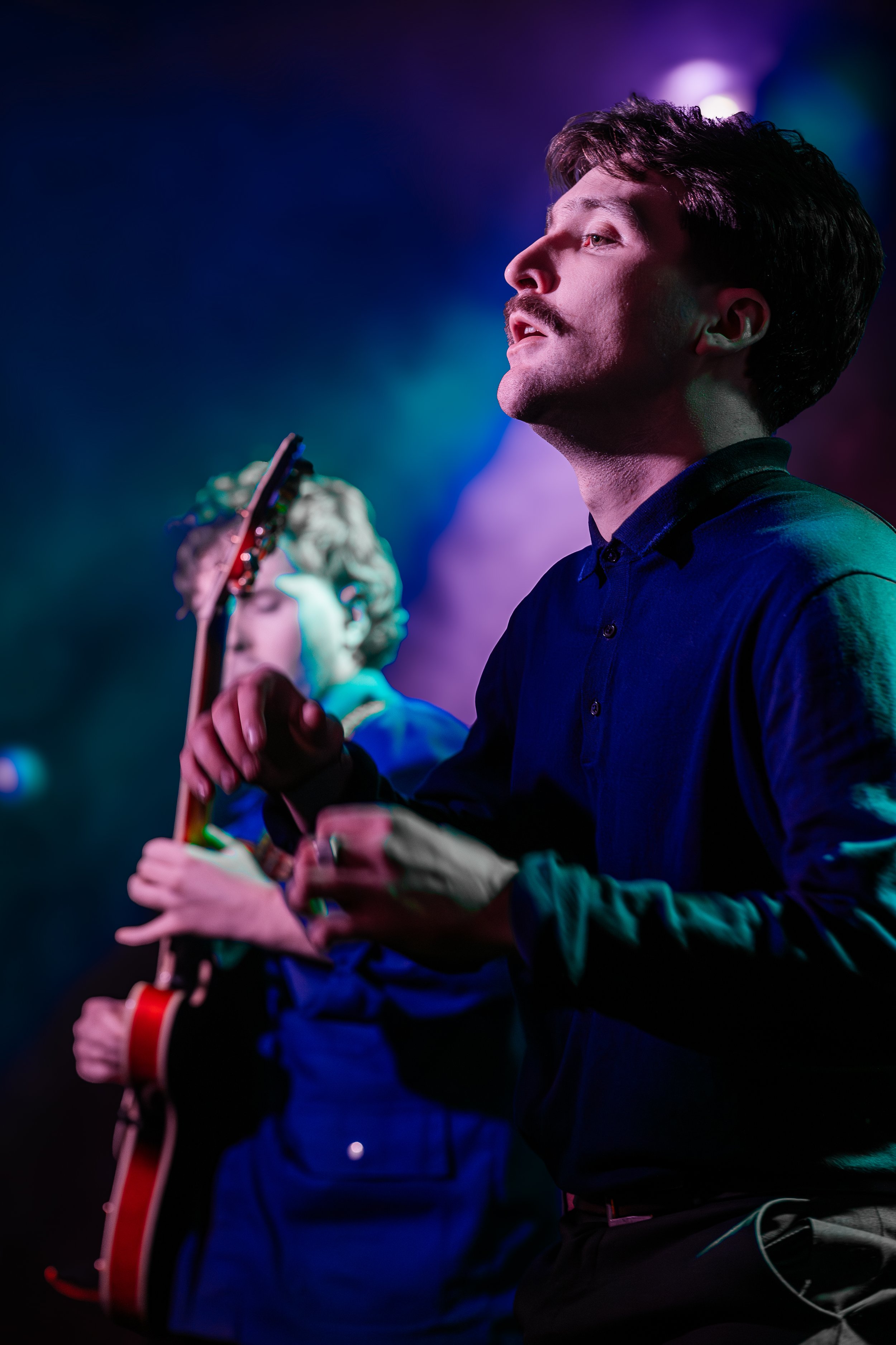 Two musicians performing on stage with colorful lighting, one playing an electric guitar and the other singing.