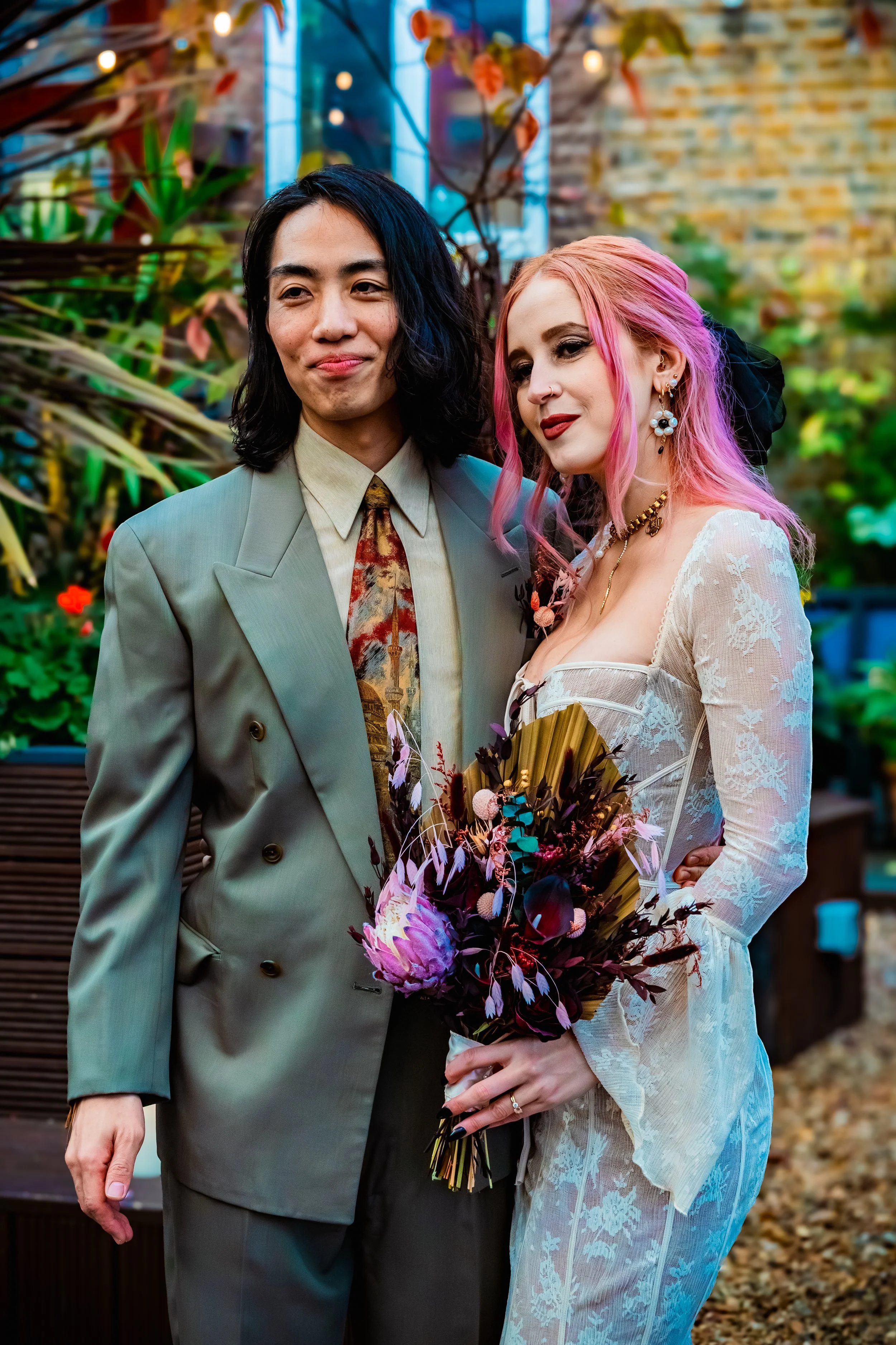 A young man in a grey suit and a young woman with pink hair in a white lace dress holding a bouquet of flowers, standing outdoors among plants and colorful lights.